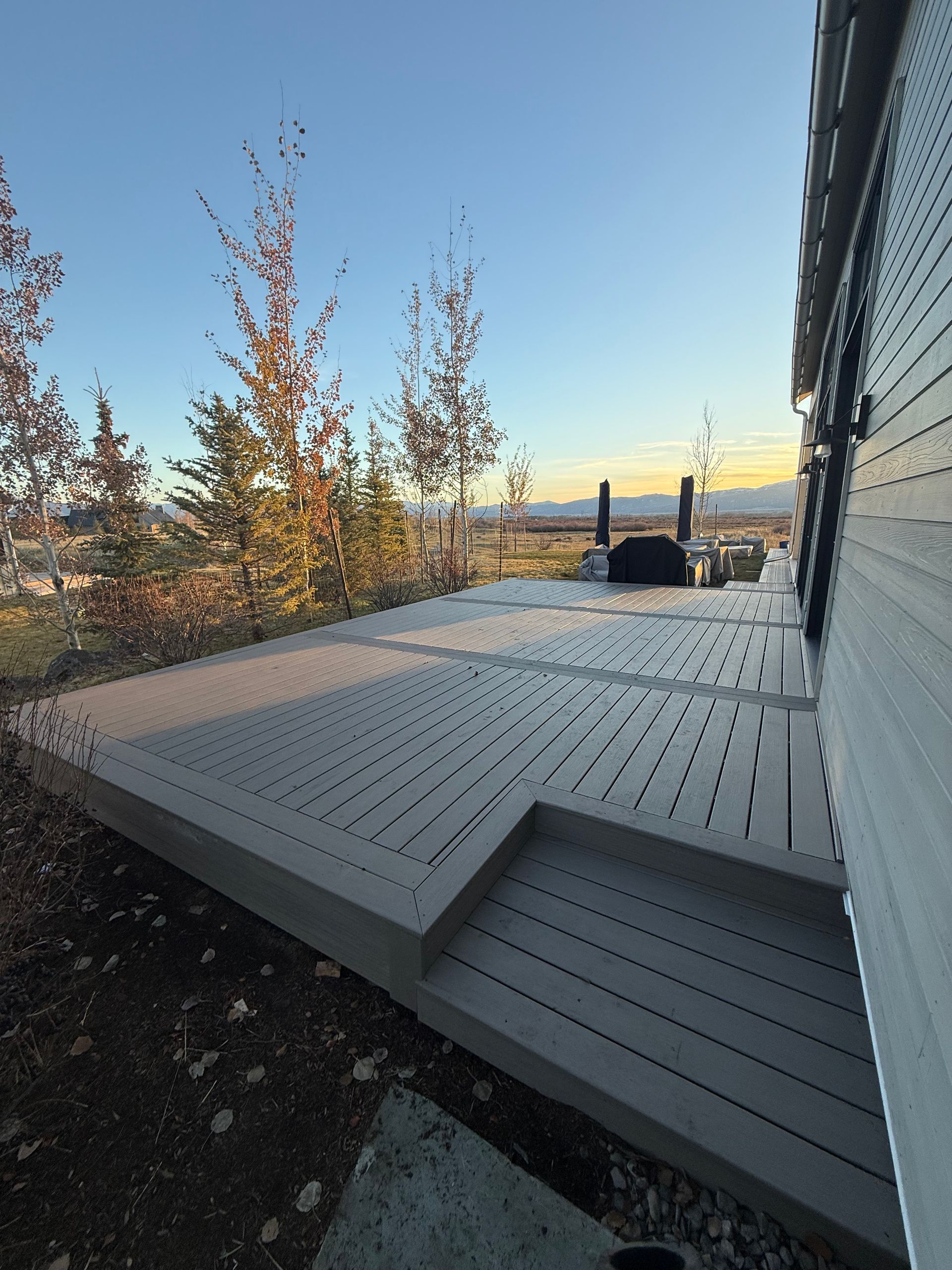 White deck with low steps beside a house at sunset, with trees and distant water in view