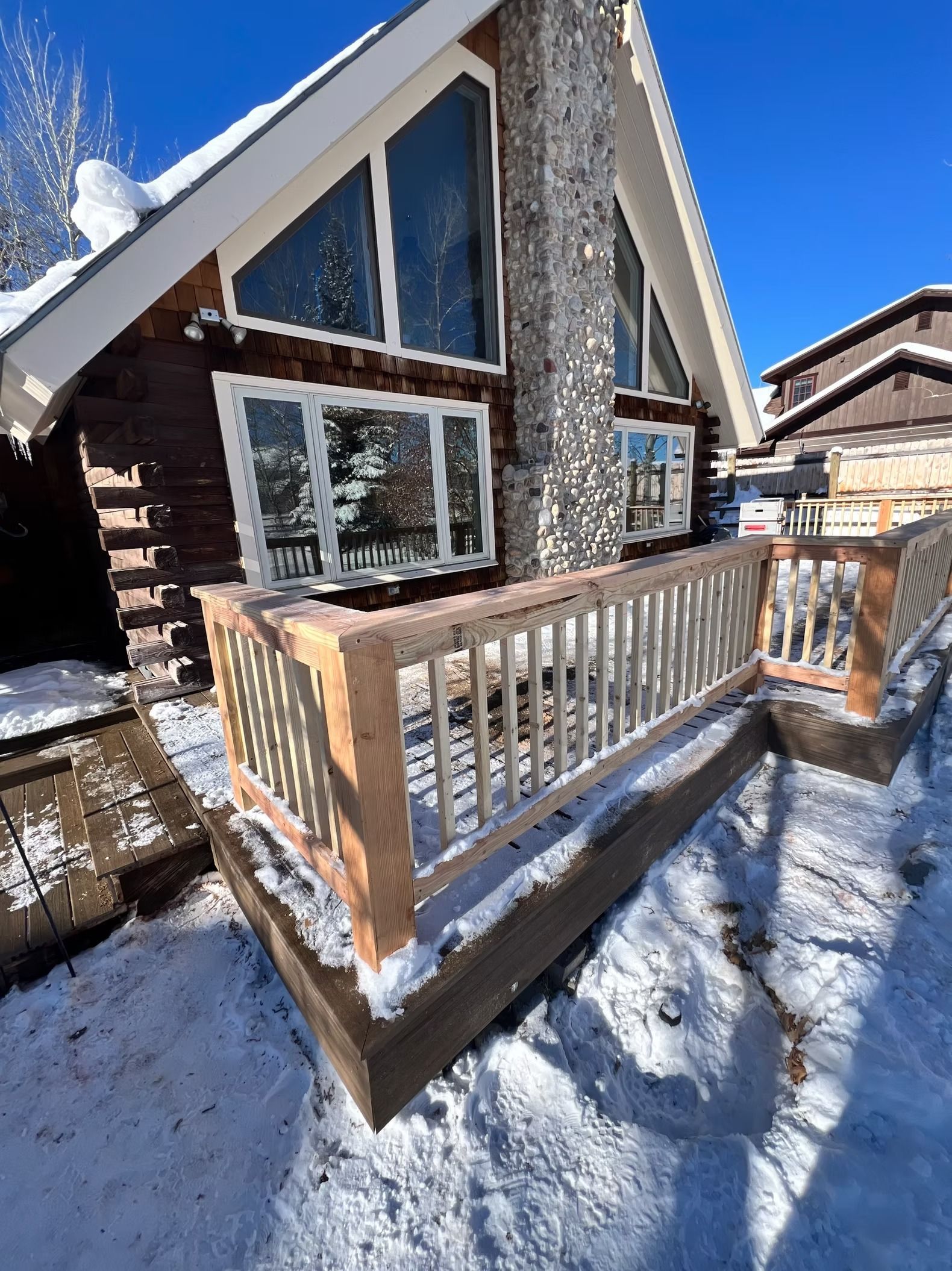 Snowy cabin deck with wooden railing outside a steep-roof house under a bright blue sky