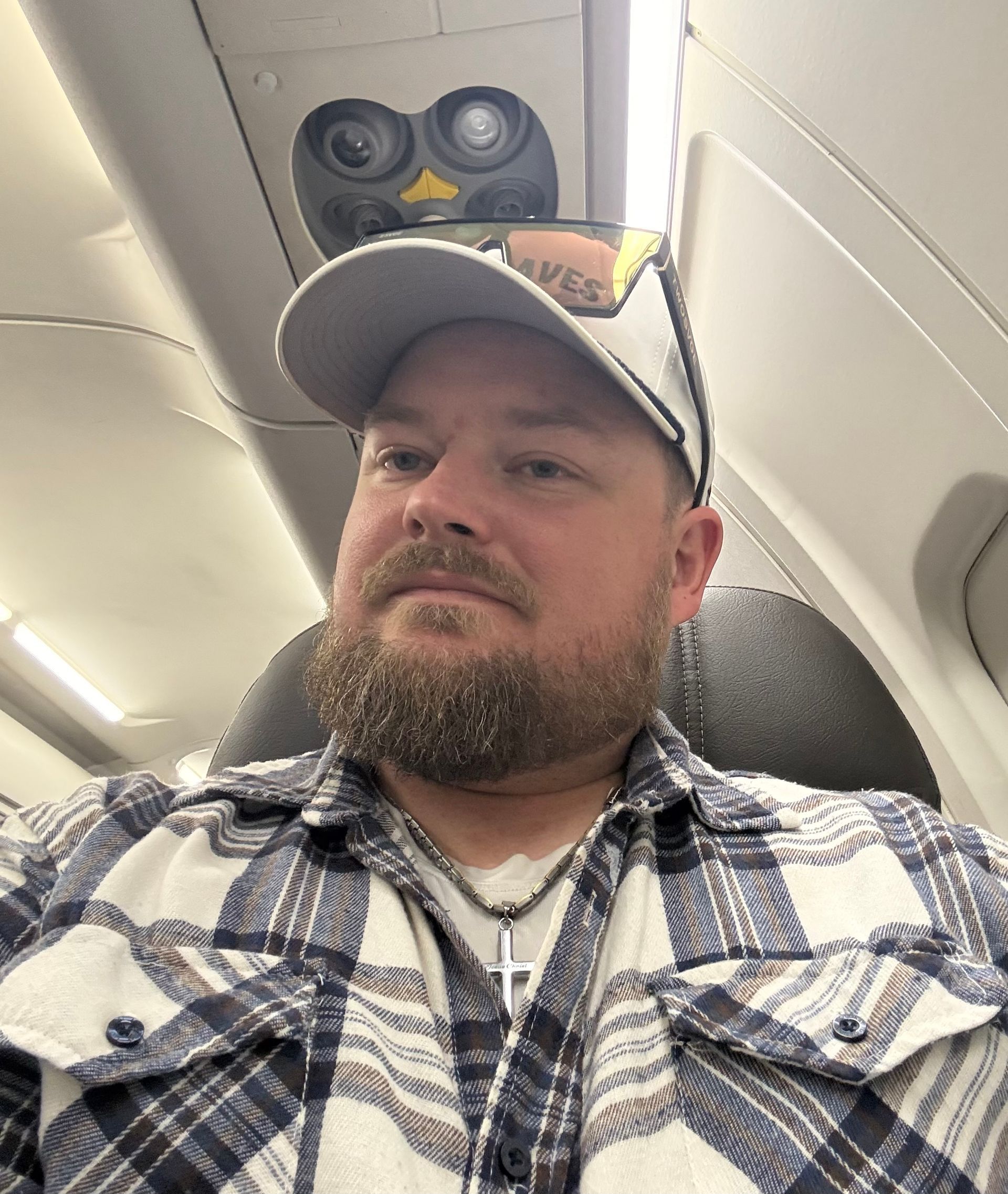 Man wearing a baseball cap and plaid shirt seated on an airplane, looking to the side.
