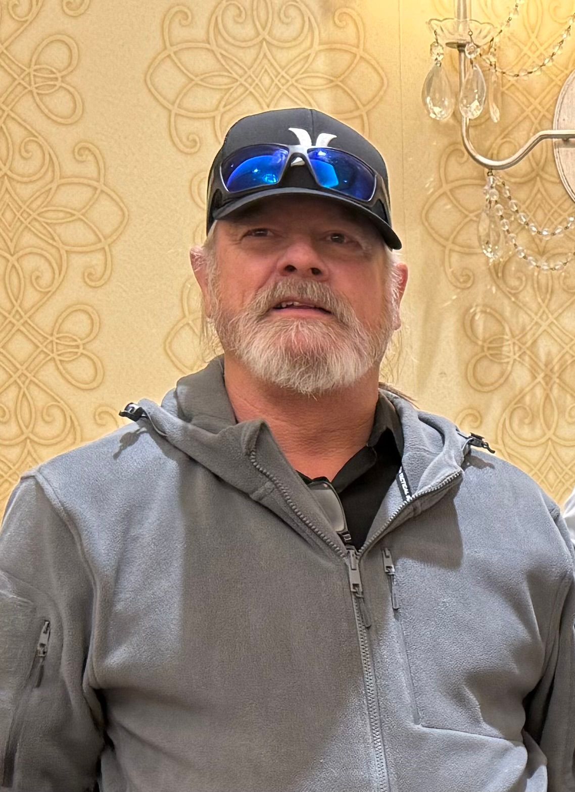 Man in a gray hoodie and black cap standing indoors against a patterned wall