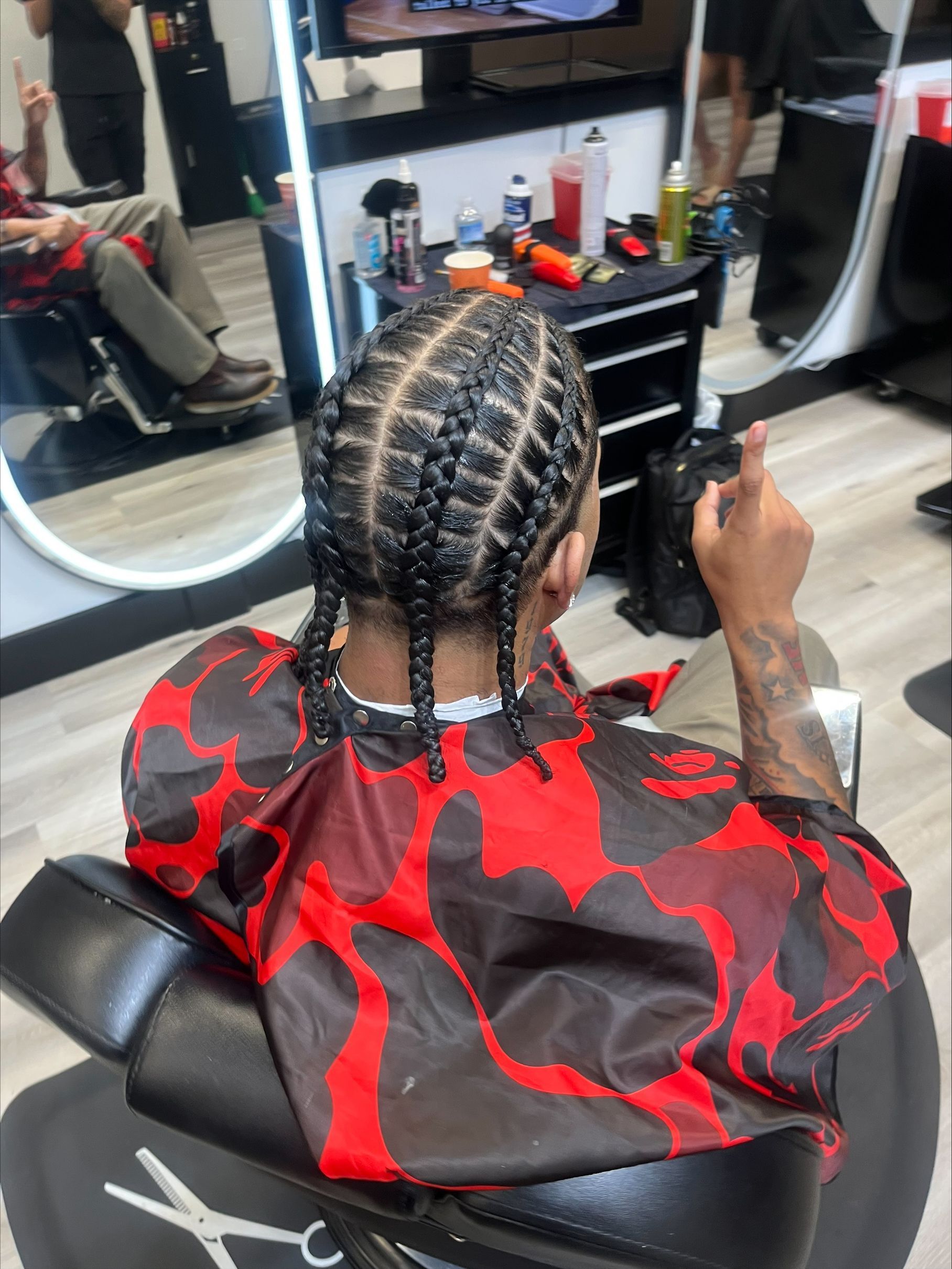 Person with braided hair and red cape sitting in a barber chair, using a phone in a salon mirror view