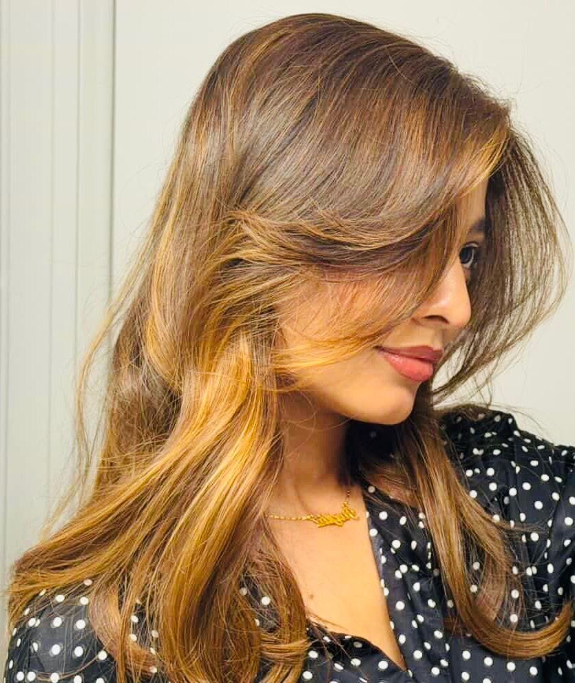 Woman with long, layered brown hair, wearing a black and white polka dot top, looking right with a slight smile.