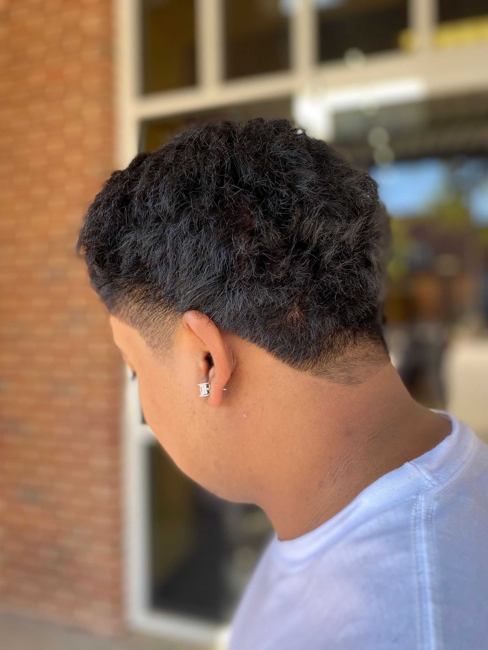 Person with a short, textured hairstyle, wearing a white shirt, against a brick wall.