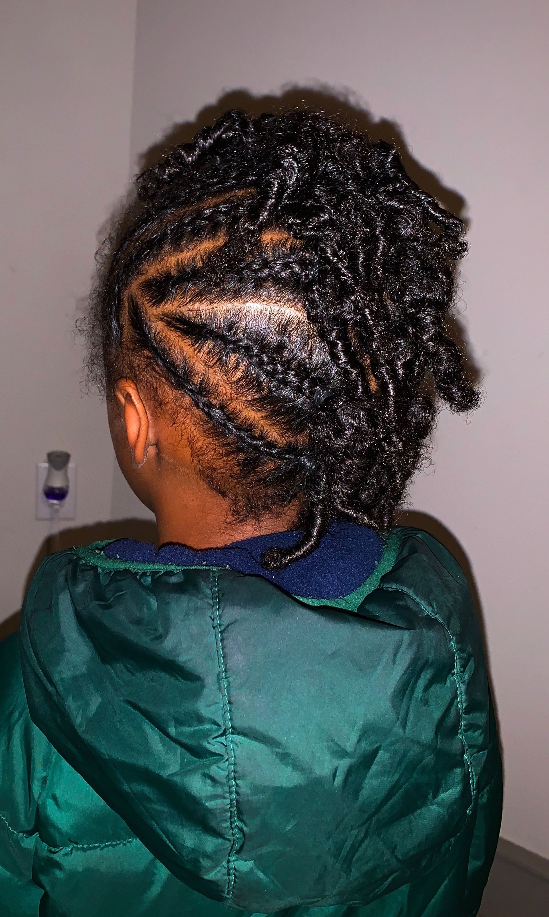 Person with dark, tightly curled hair in a side-swept style. Wearing a green jacket.
