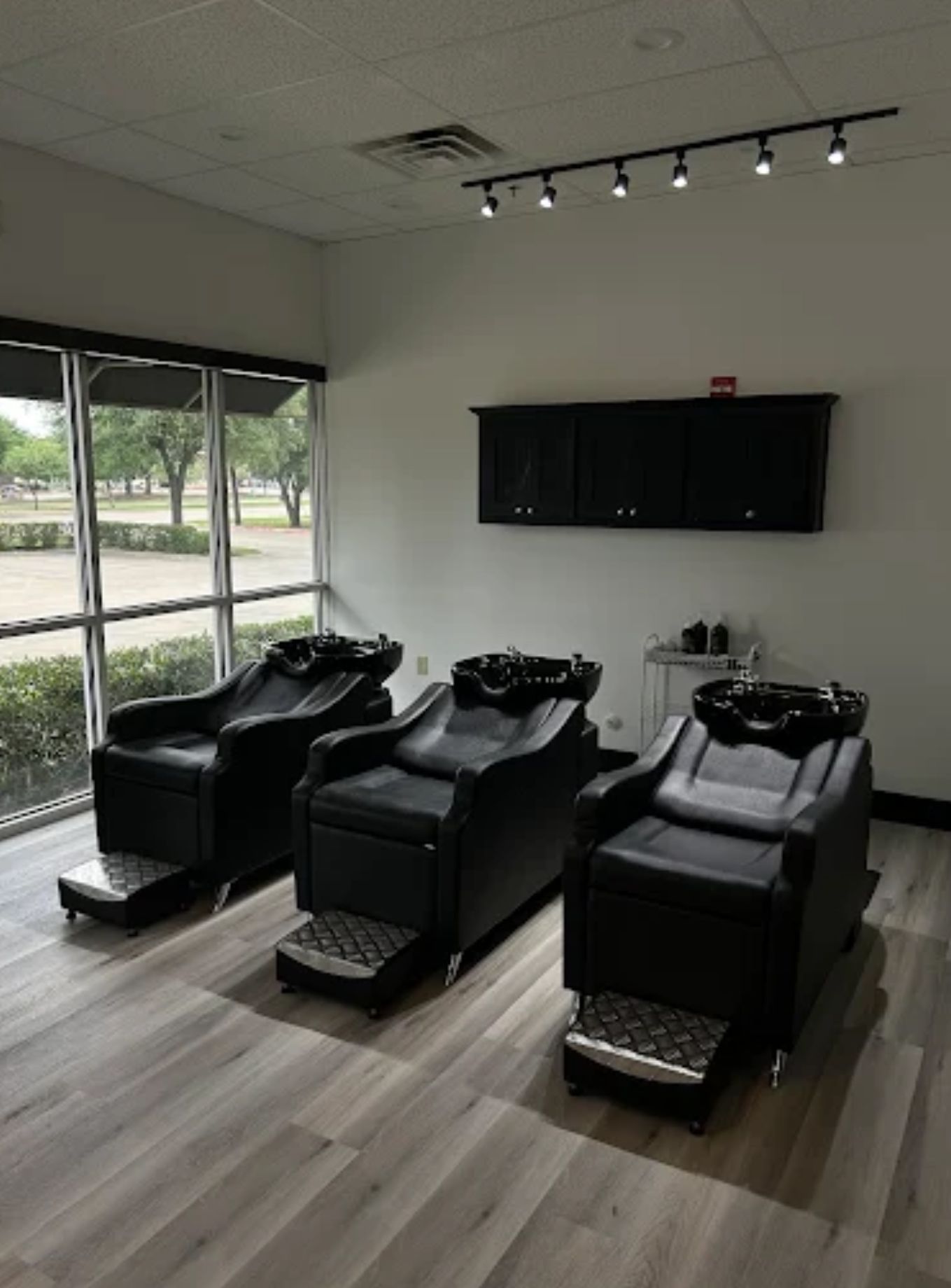Three black salon chairs with sinks, under a black cabinet, near a window.