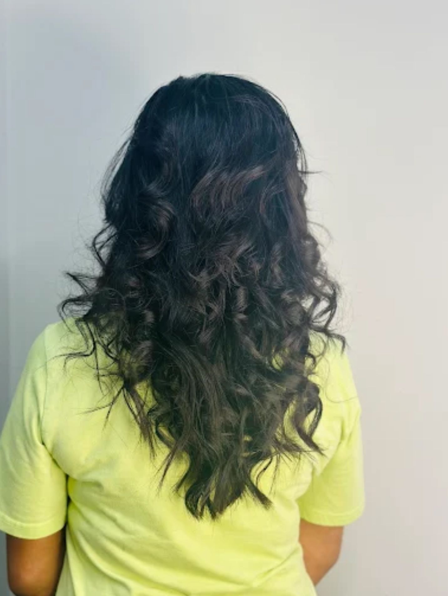 Woman with wavy black hair, wearing a light yellow shirt, facing away from the camera.