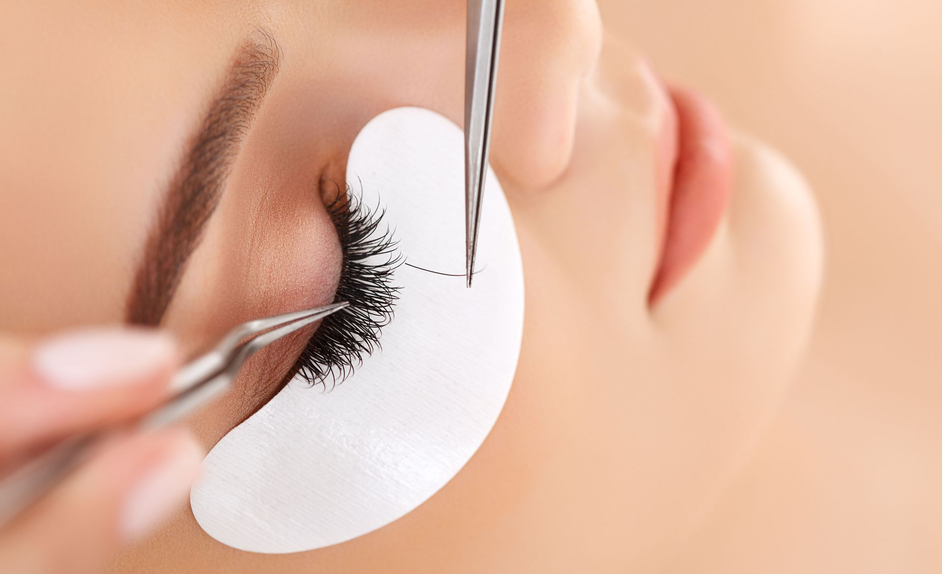 Close-up of eyelash extensions being applied with tweezers and a protective under-eye pad