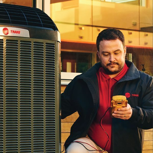 HVAC technician in red shirt checks AC unit with a yellow meter; outdoor setting.