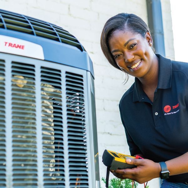 Woman smiling, holding a multimeter, next to a Trane air conditioning unit outdoors.