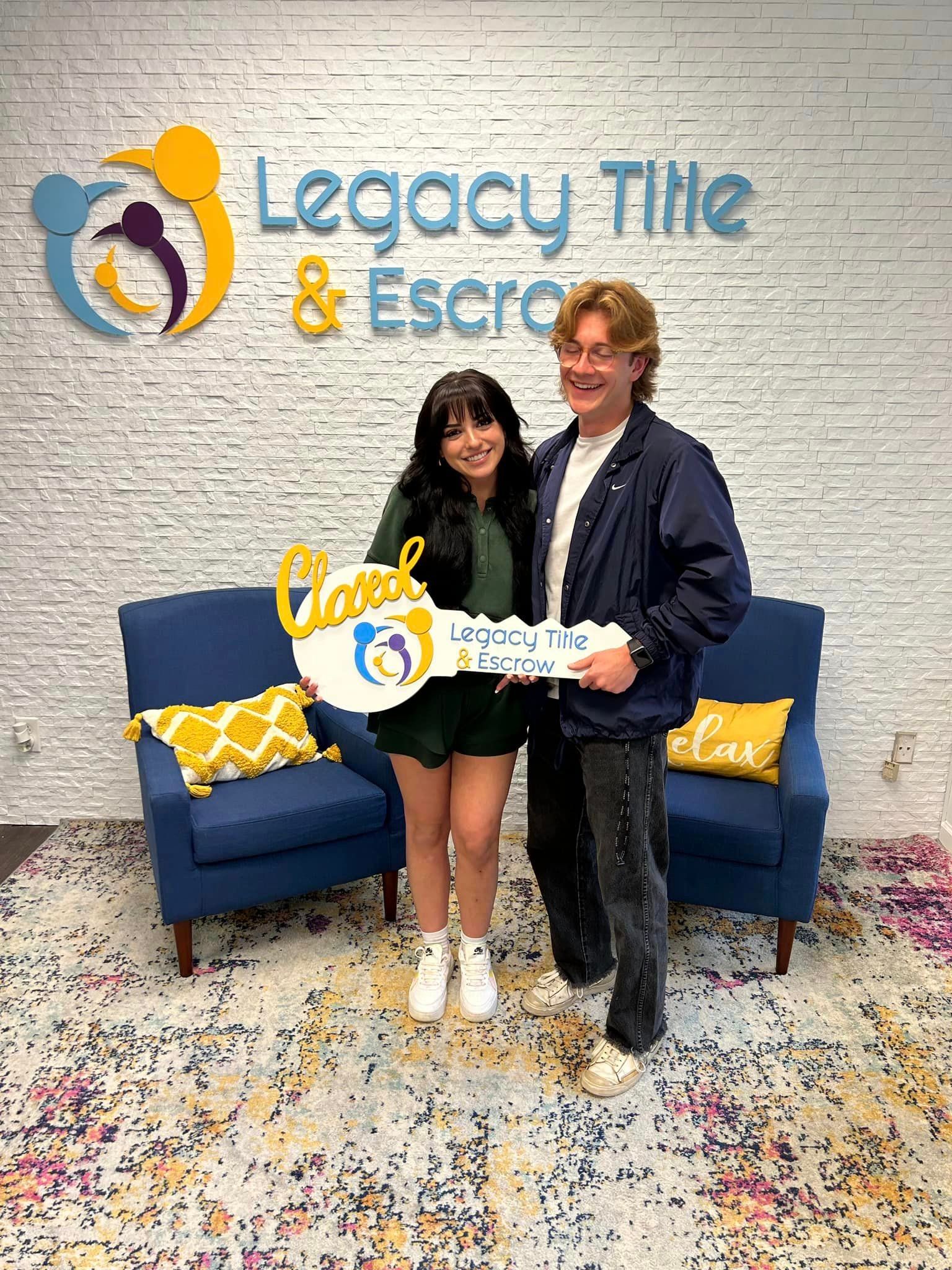 A man and a woman are holding a key in front of a legacy title and escrow sign.