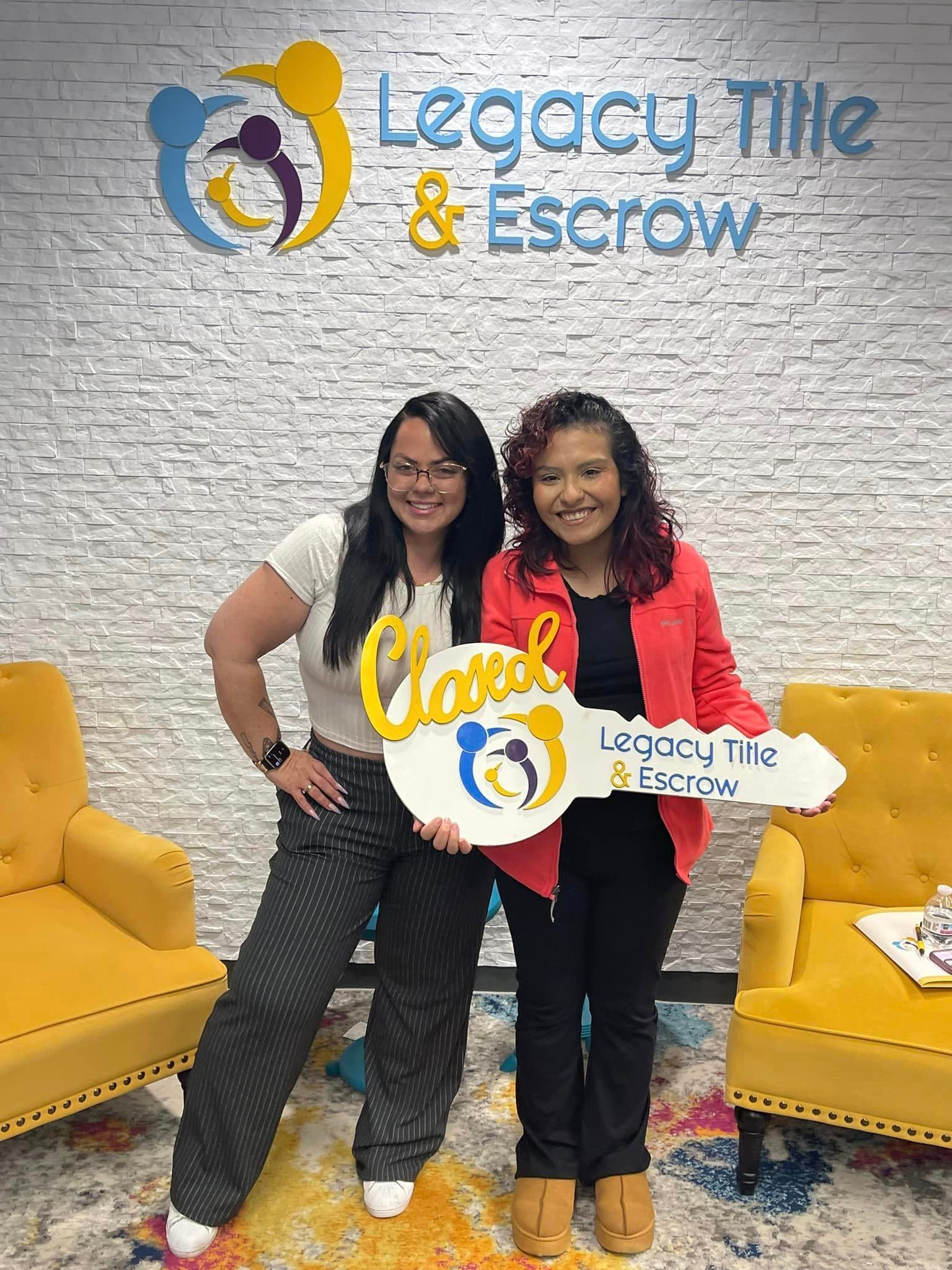 Two women are standing next to each other holding keys in front of a legacy title and escrow sign.