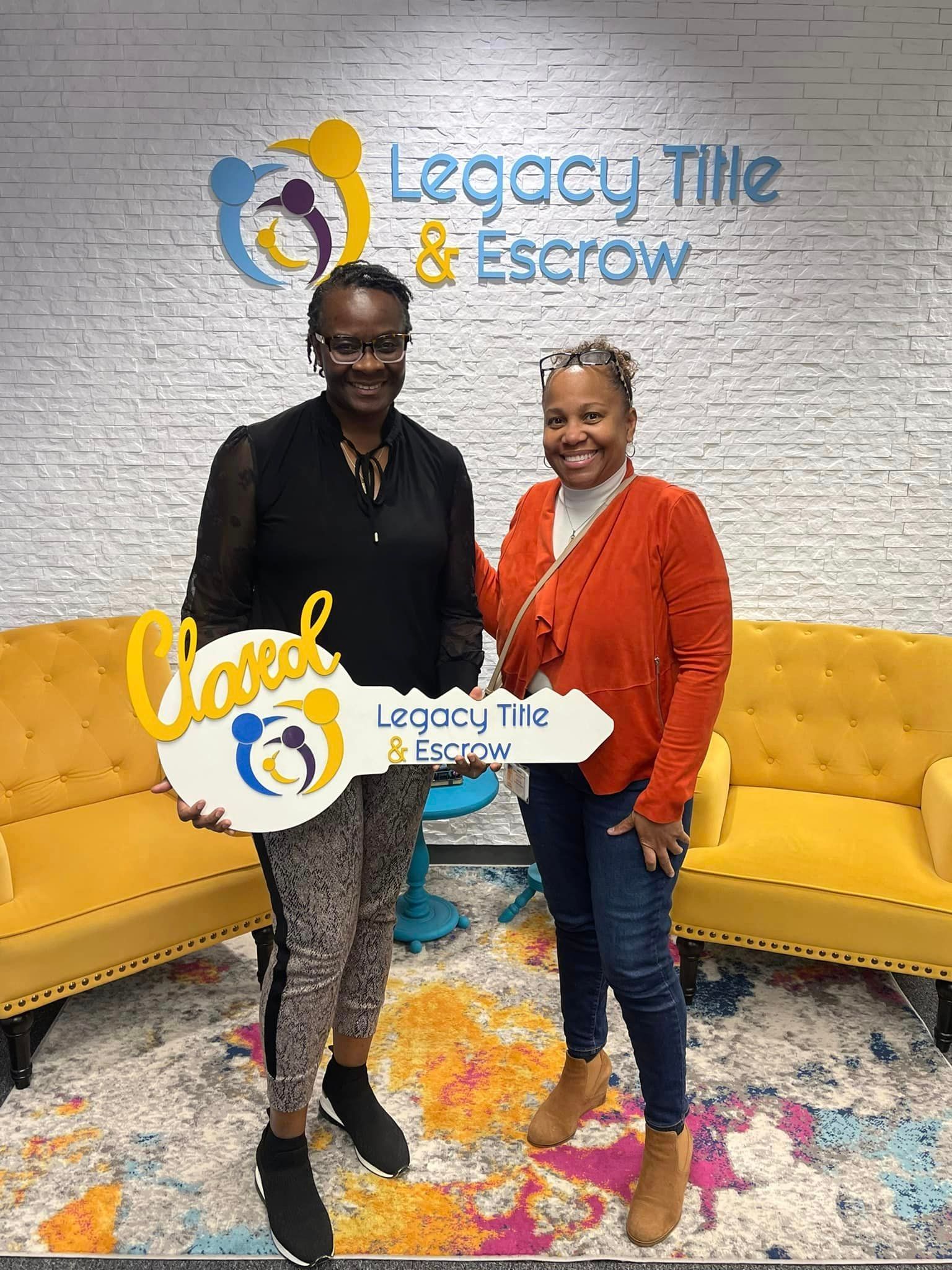 Two women are standing next to each other in front of a yellow couch holding a key.