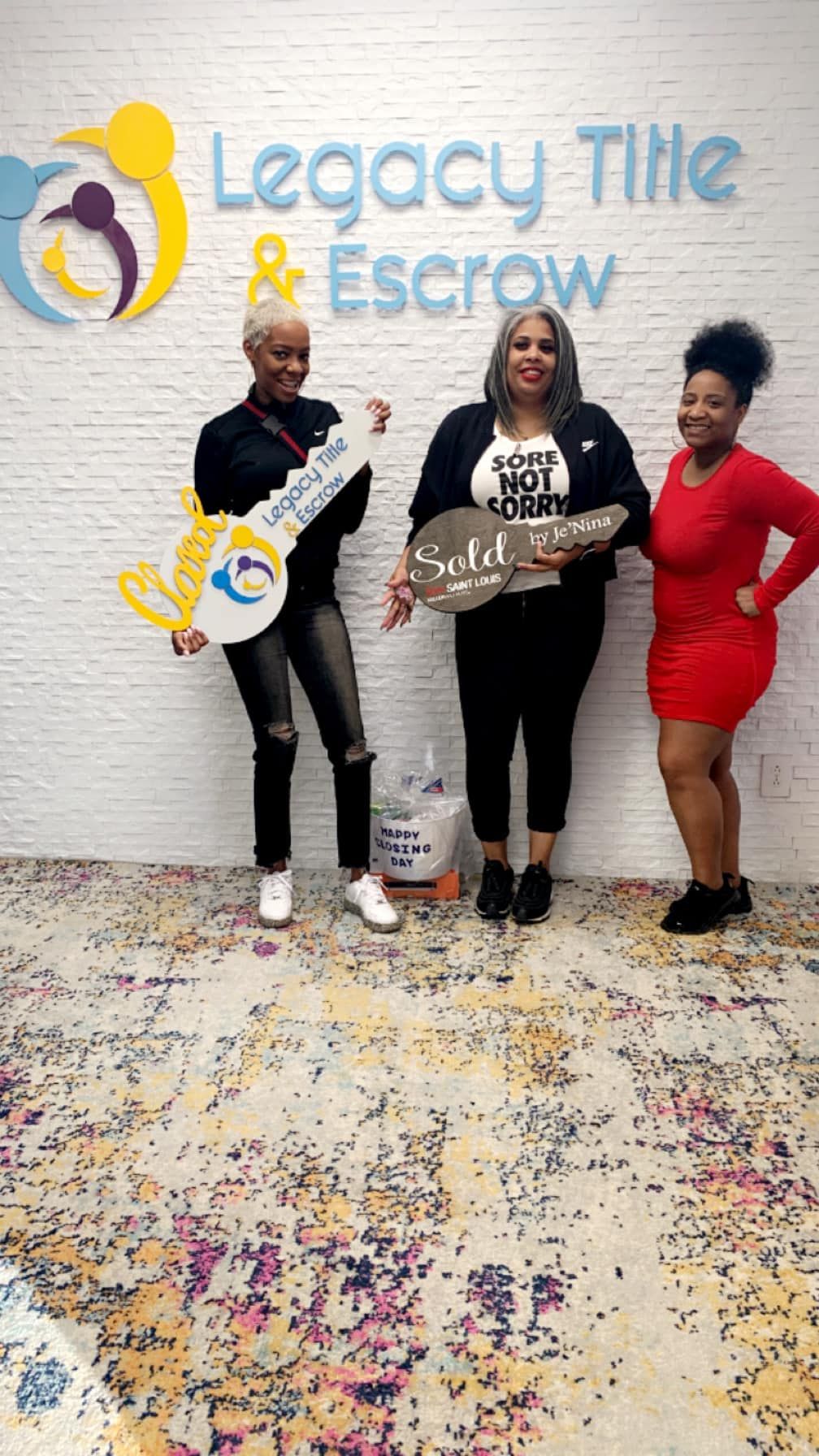 Three women are standing next to each other holding keys in front of a legacy title and escrow sign.