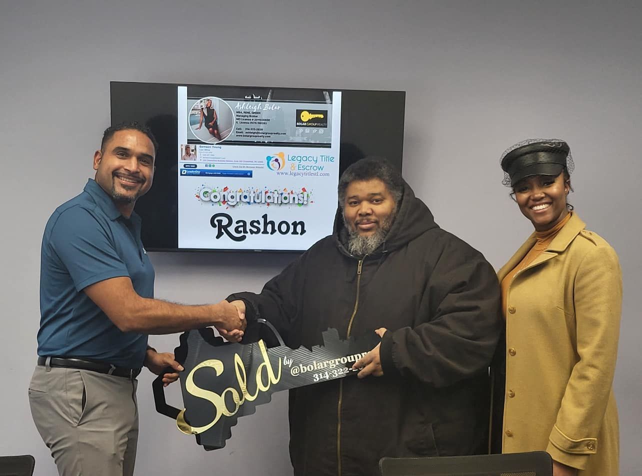 A man and a woman are shaking hands with a man holding a sold sign.