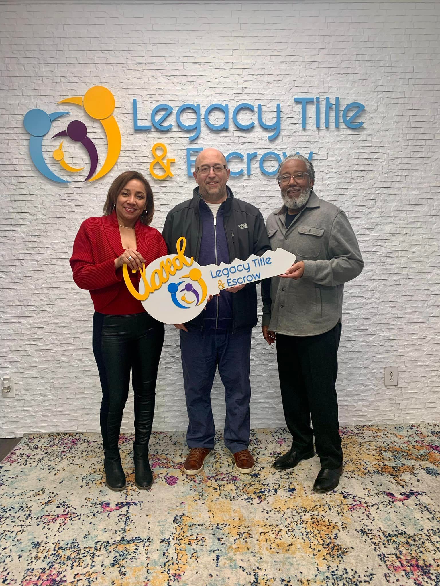 Three people are standing next to each other in front of a brick wall holding a large key.