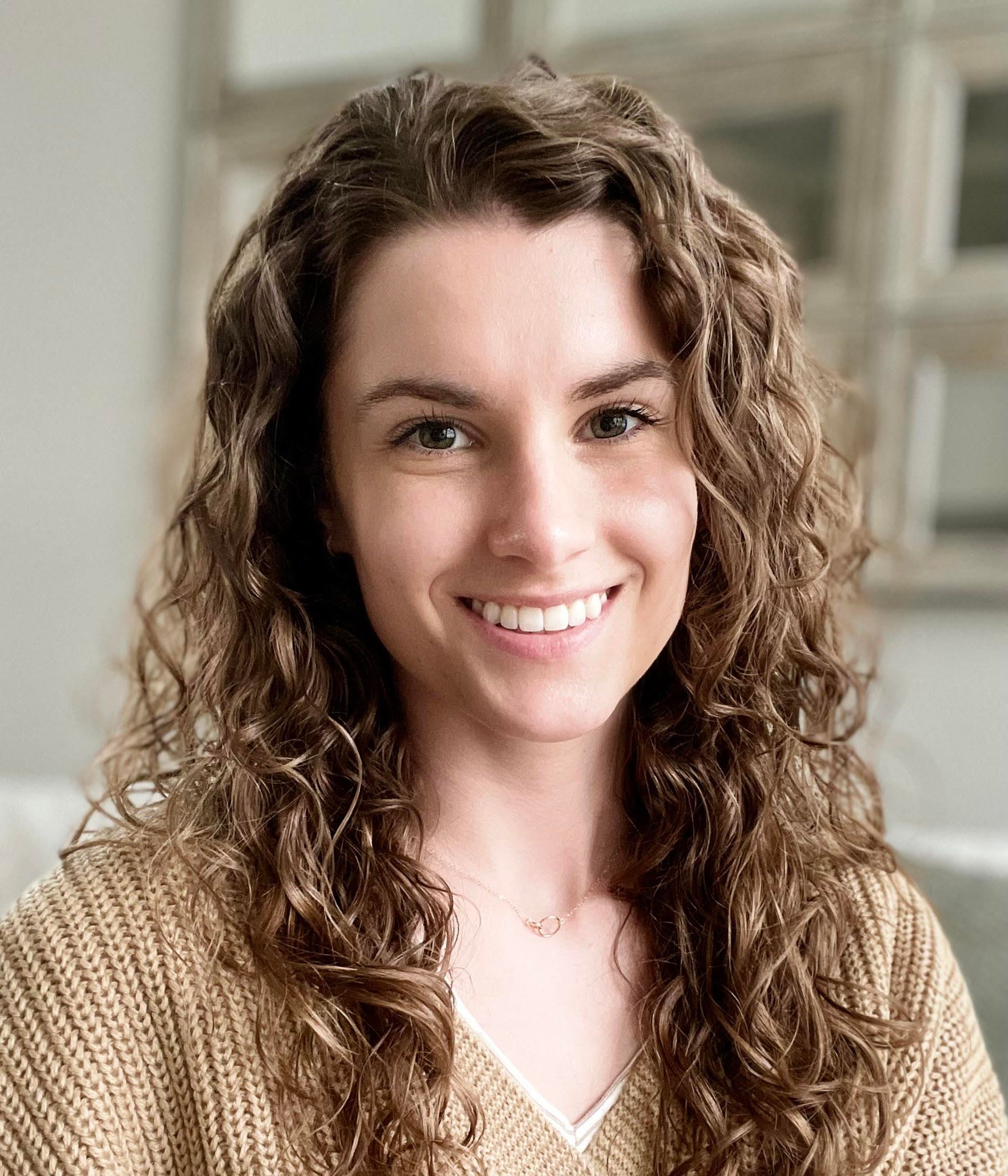 A woman with curly hair is smiling for the camera while wearing a tan sweater.