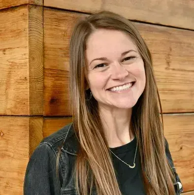 A woman with long hair is smiling in front of a wooden wall.