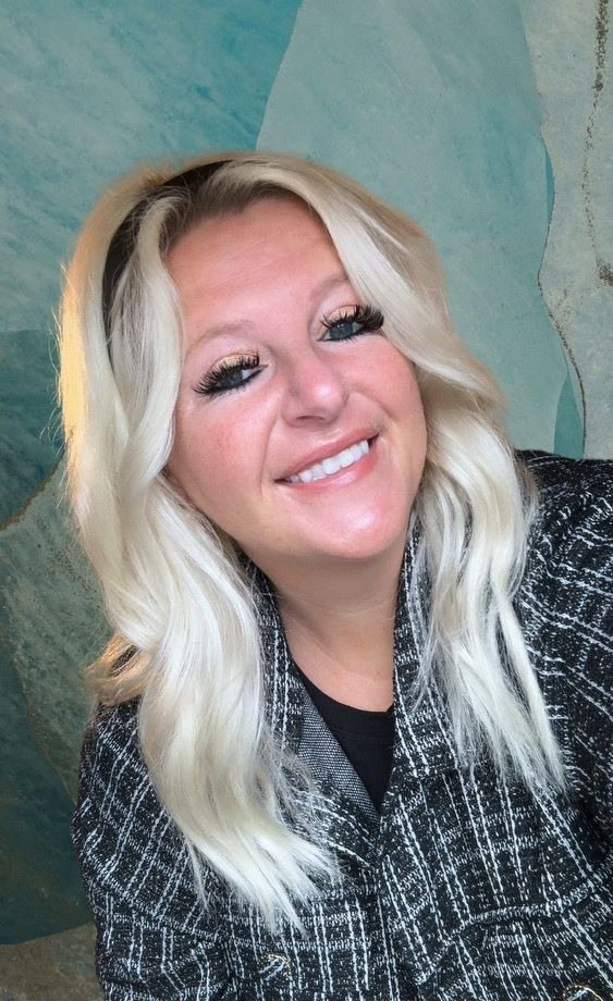 A woman with blonde hair is smiling and wearing a black and white jacket.
