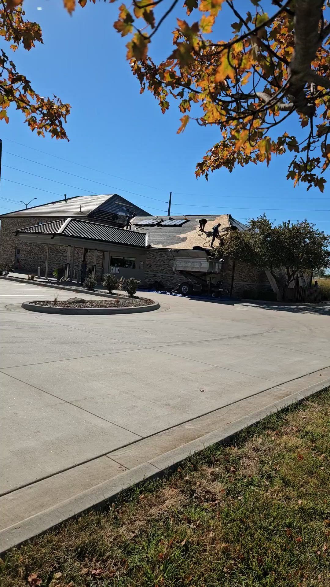Damaged building with exposed roof, possibly from fire. Paved driveway in front with autumn foliage overhead under a blue sky.