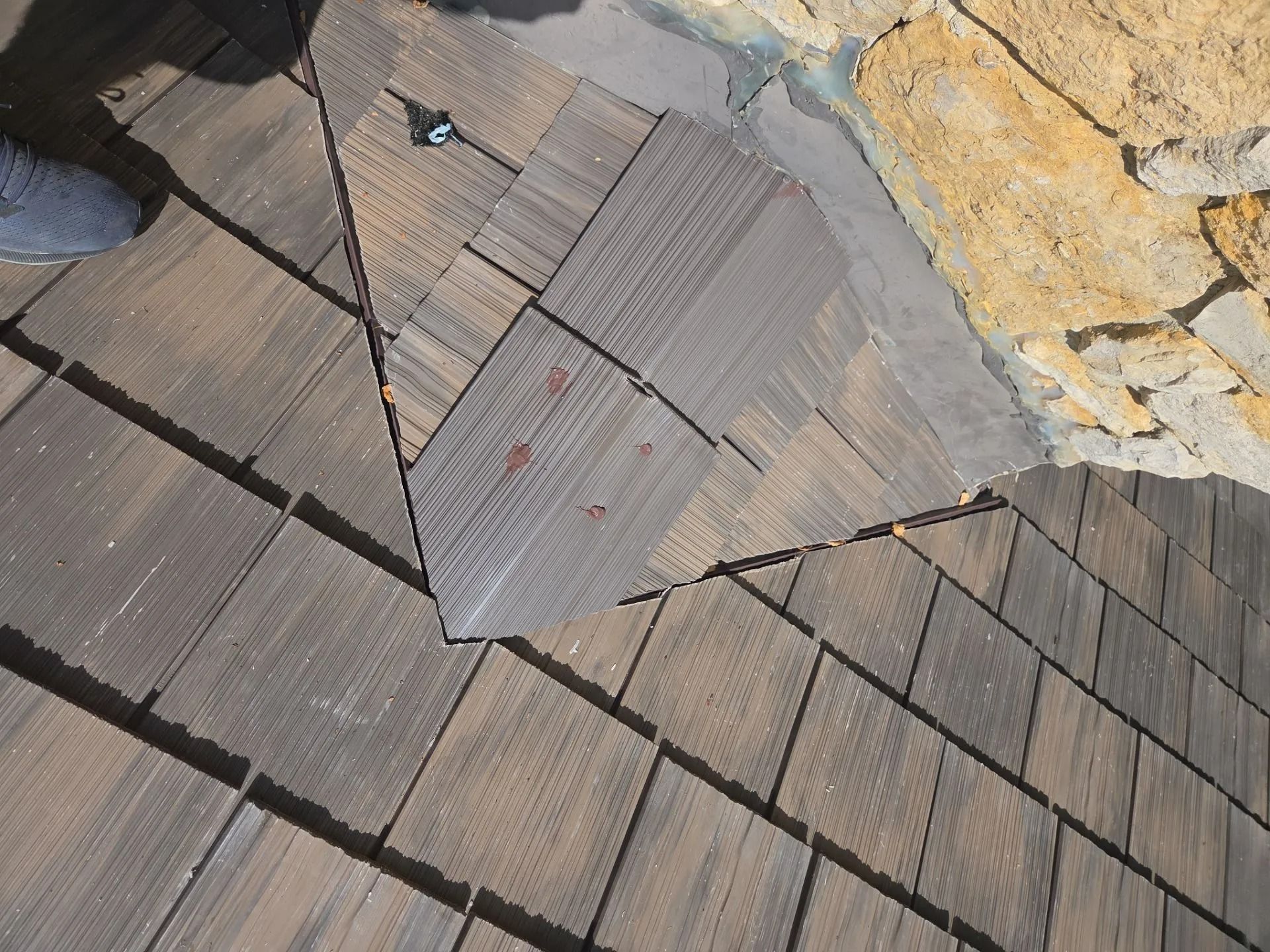 Close-up of a damaged roof with brown composite shingles adjacent to a stone wall. Some shingles are missing.