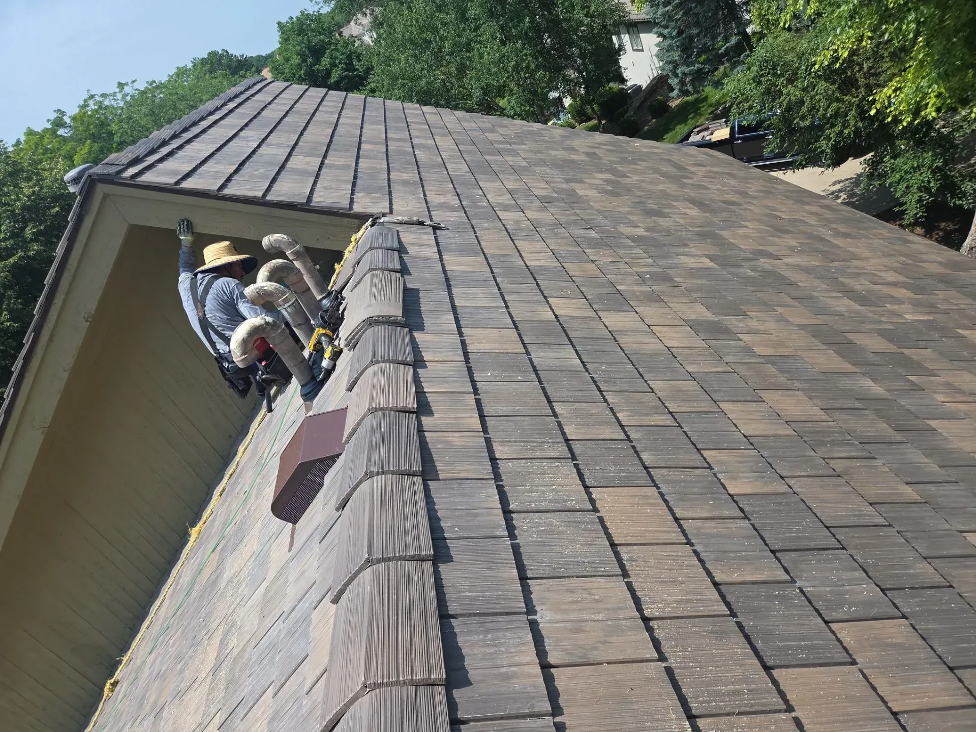 Two workers on a brown shingled roof, one near the edge, securing something. Yellow safety gear.