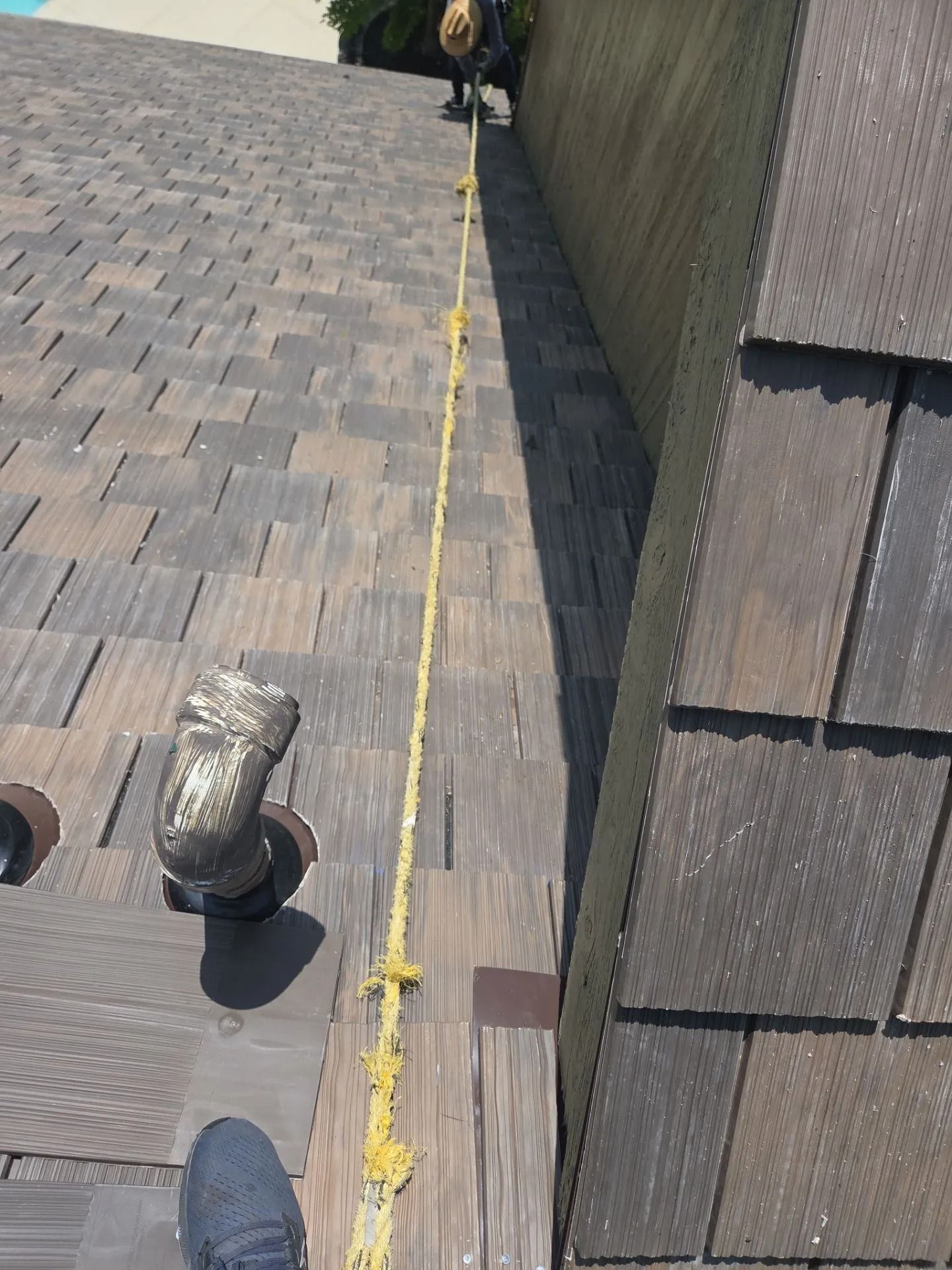 Roofer on a shingled roof with safety line, near a vent pipe. Brown roof tiles and the side of a building are visible.