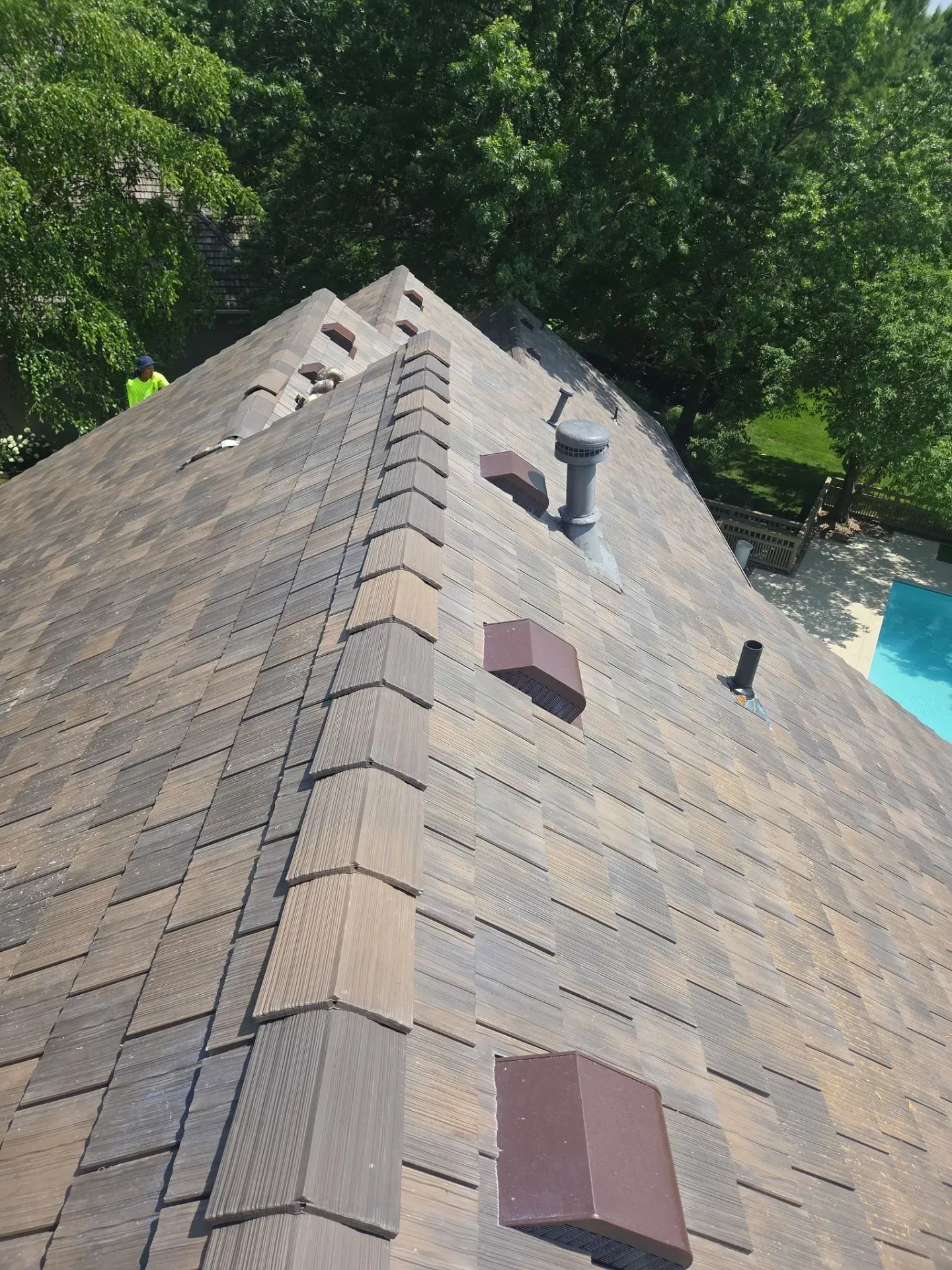 Roof with brown and gray shingles; a vent pipe and two small, brown covers visible. Green trees and a pool are in the background.