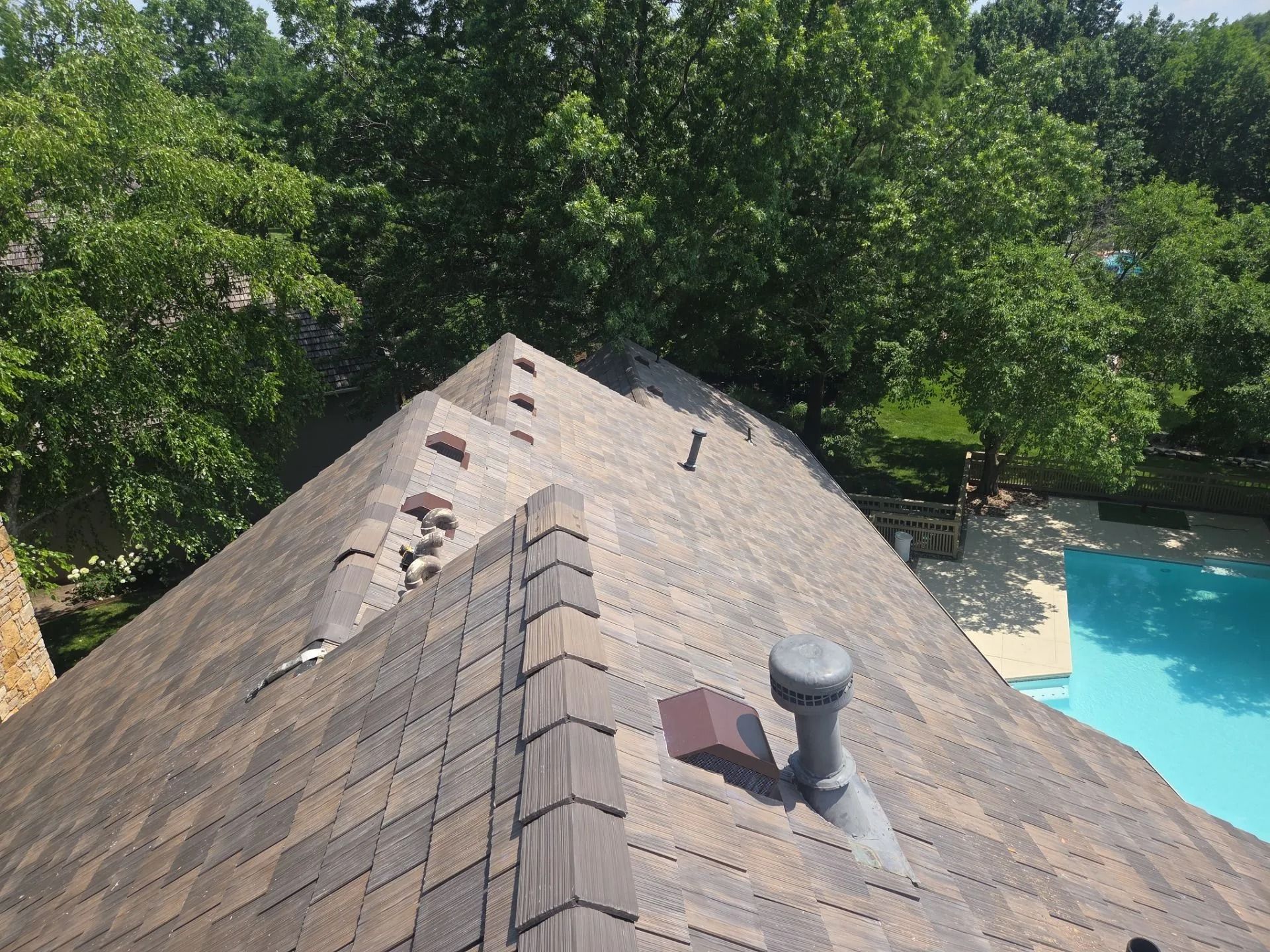 Overhead view of a roof with brown shingles, a vent, and green trees with a pool in the background.
