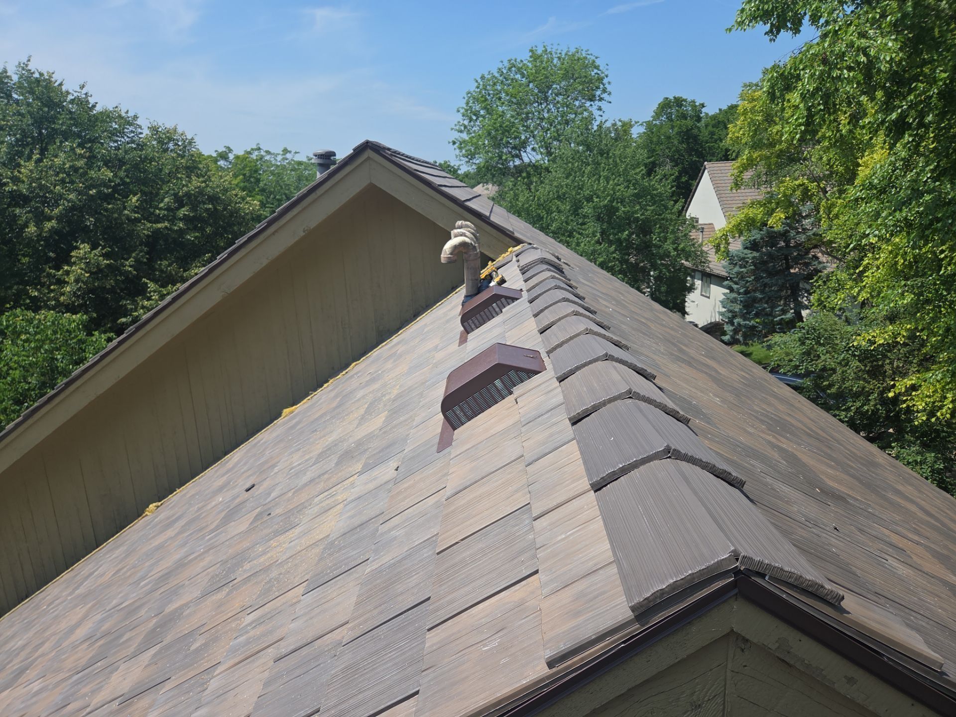 Damaged brown shingle roof on a house with a missing vent, trees in background, sunny day.