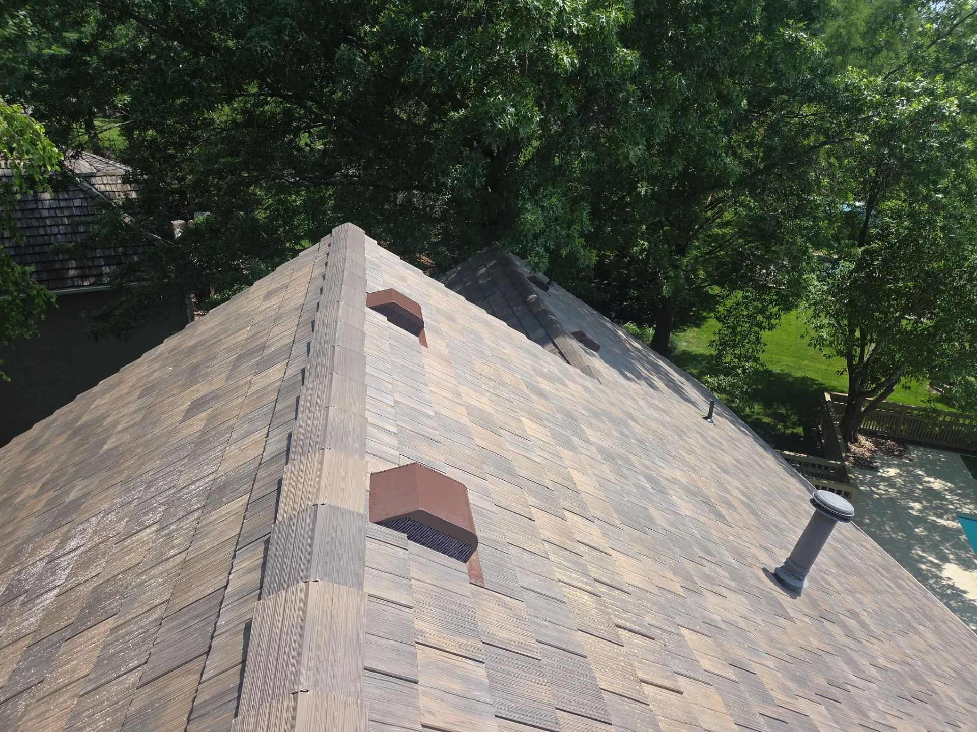 Overhead view of a house roof with faded shingles and vents. Brown roof with trees in the background.