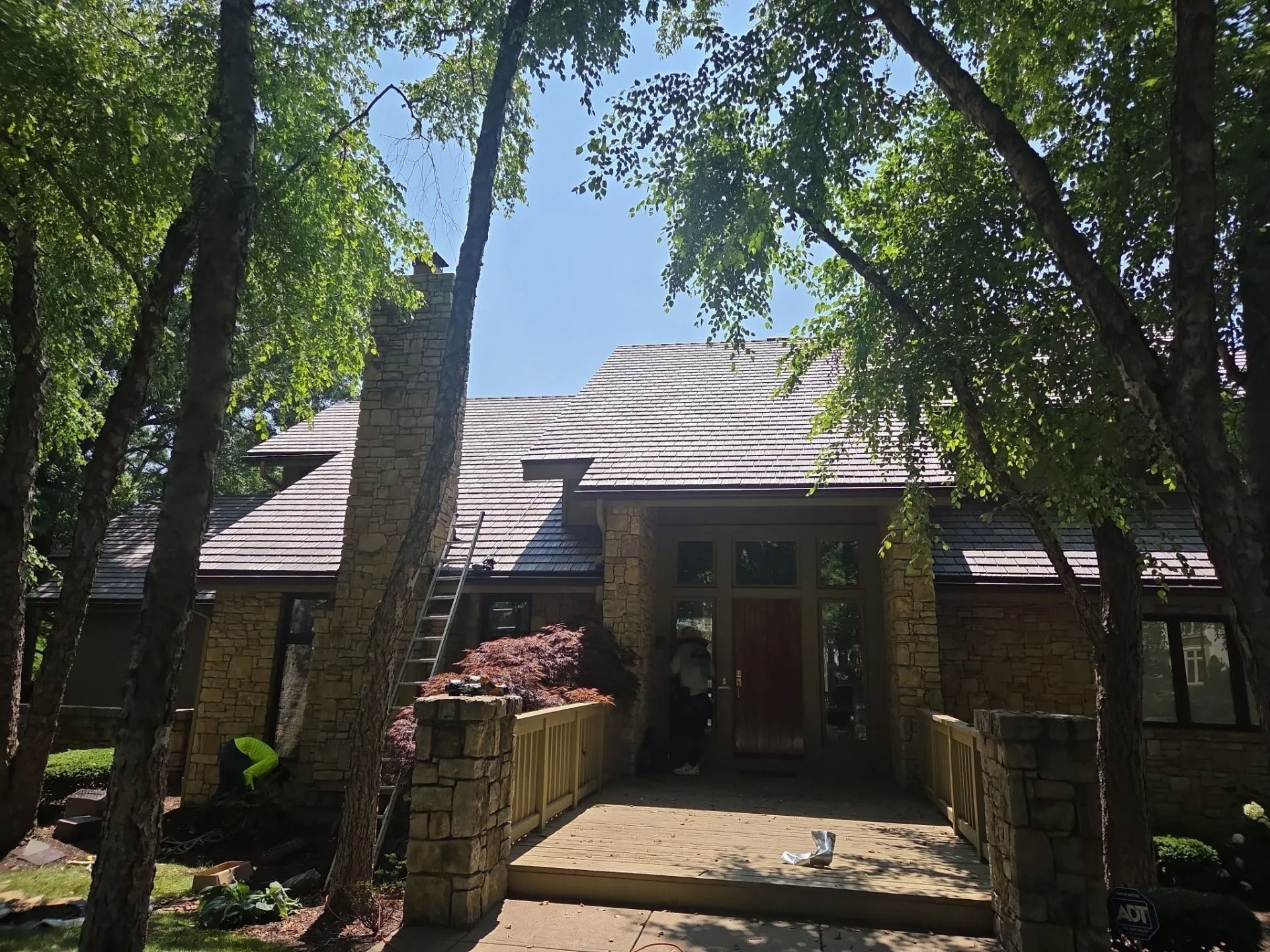 House with brown roof, stone facade, and trees. A ladder is leaning against the roof.