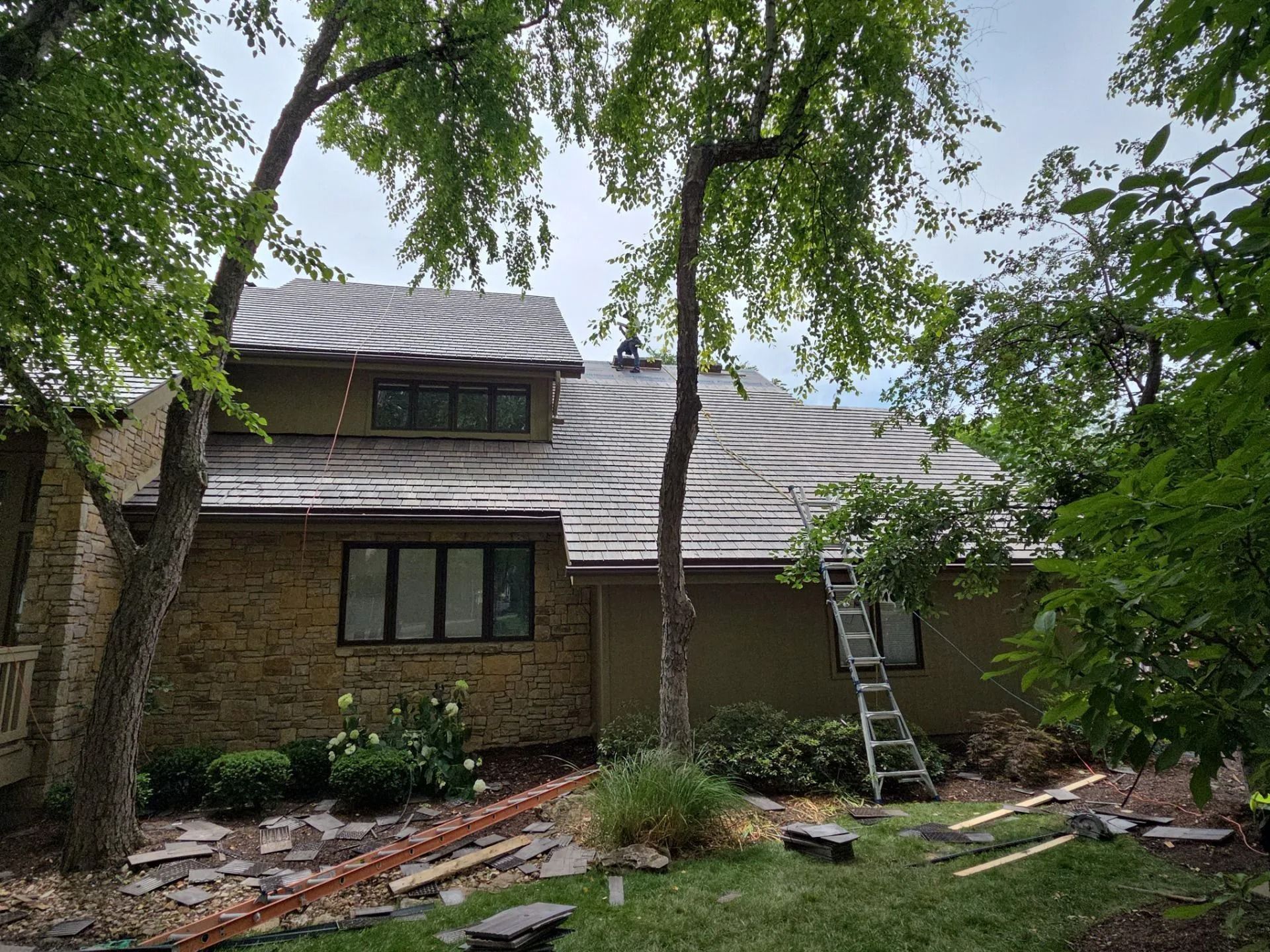 House exterior with new roof installation, ladder, and trees. Brown roof, beige brick, and green trees.