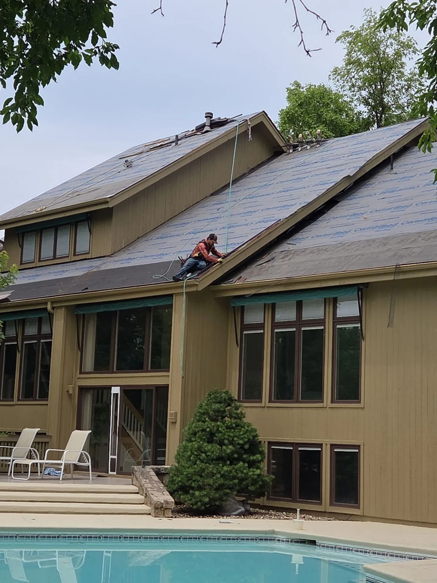 Two people working on a roof, attached to a light brown house with a pool in front.