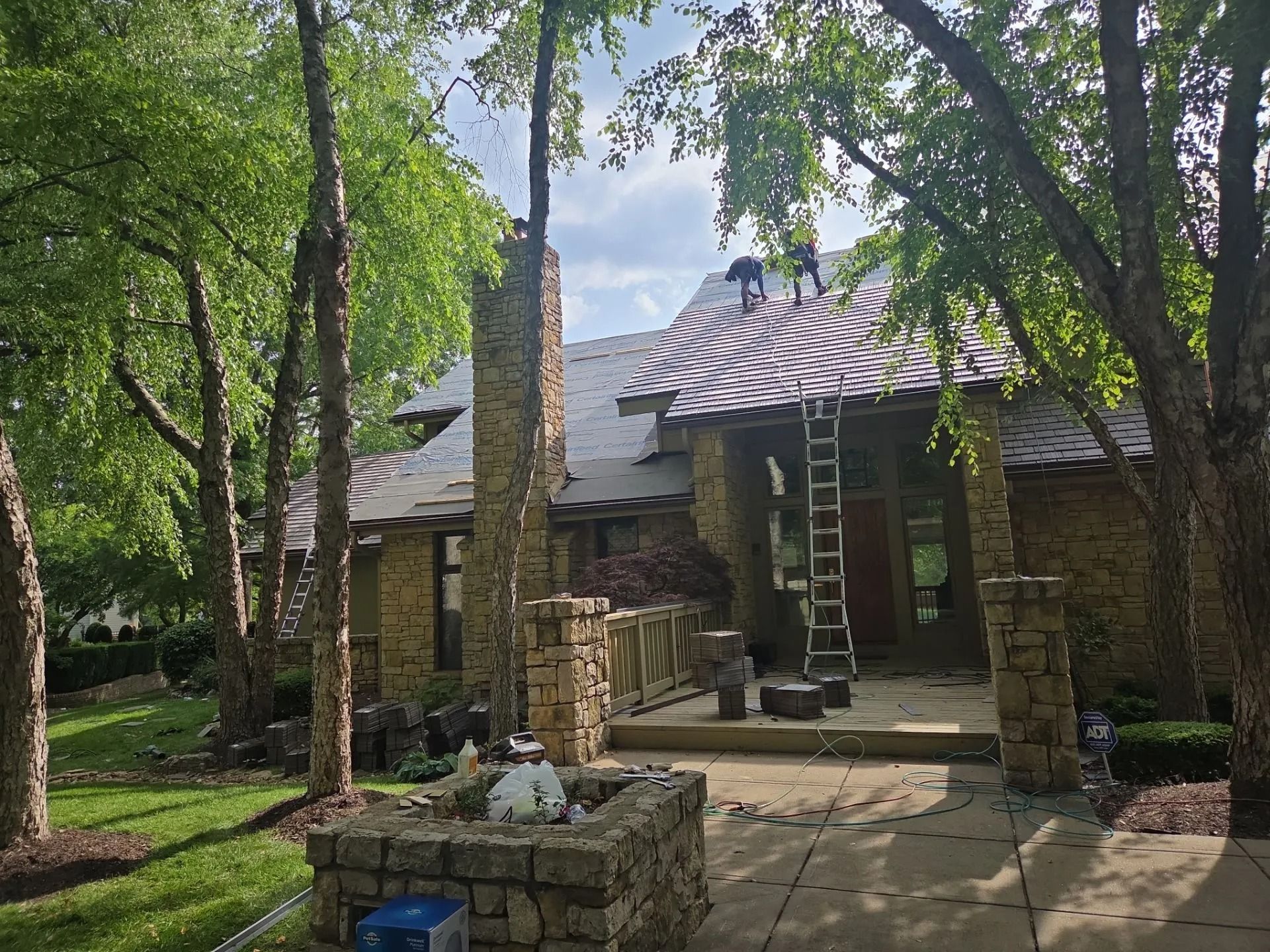 Workers replacing roof tiles on a house with stone pillars, a ladder, and surrounding trees.