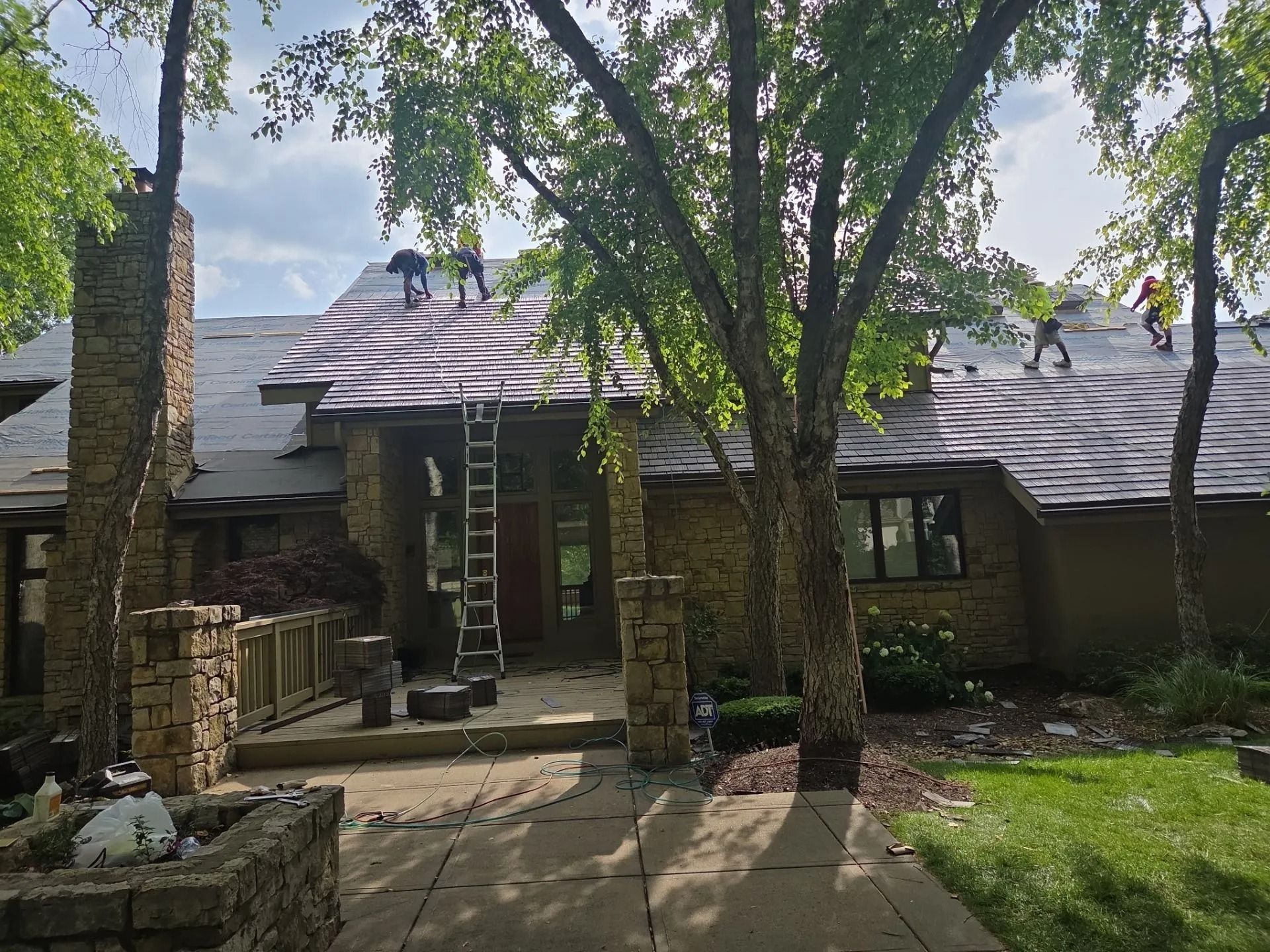 Roofers working on a house, removing old shingles. A ladder is propped against the roof.