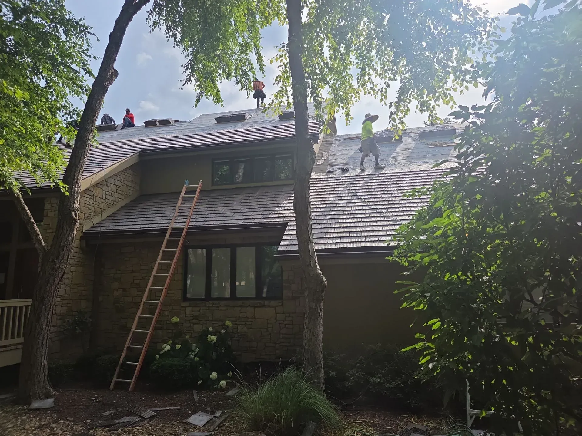 Roofers working on a house roof on a sunny day. Orange ladder against the building, trees surround.
