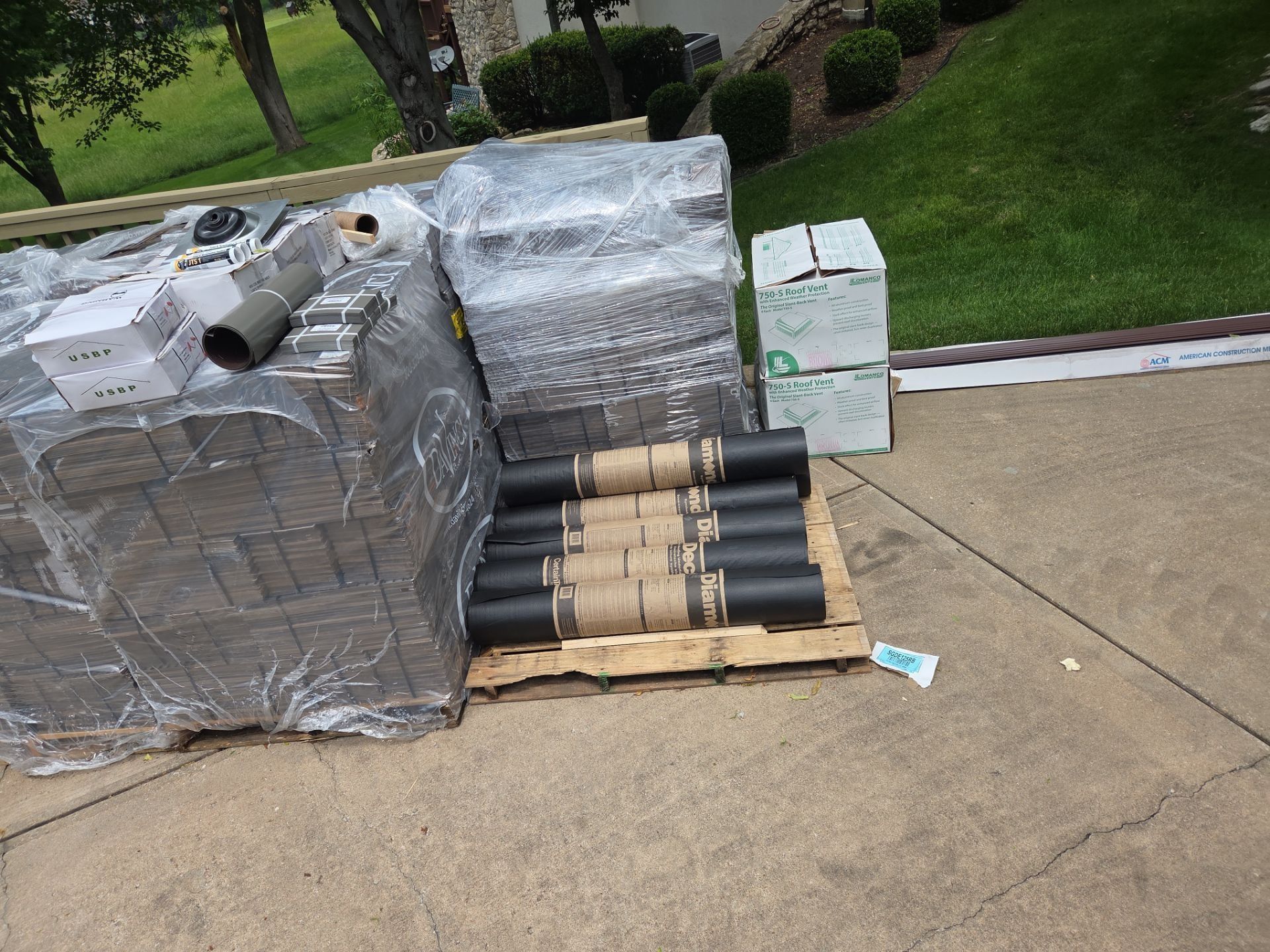Roofing materials, including shingles and rolled roofing, are on a pallet in a driveway.