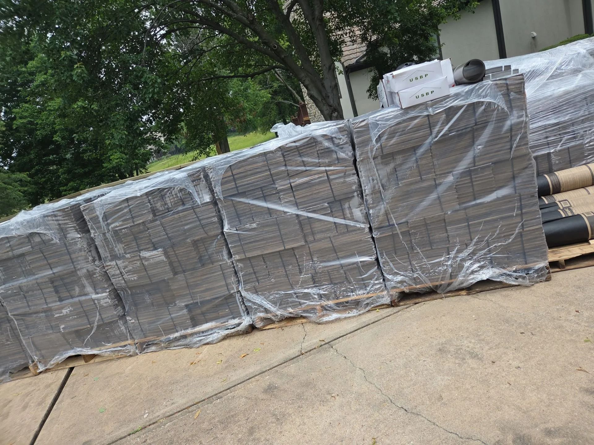 Pallet of gray paving stones wrapped in clear plastic, outdoors near a house.