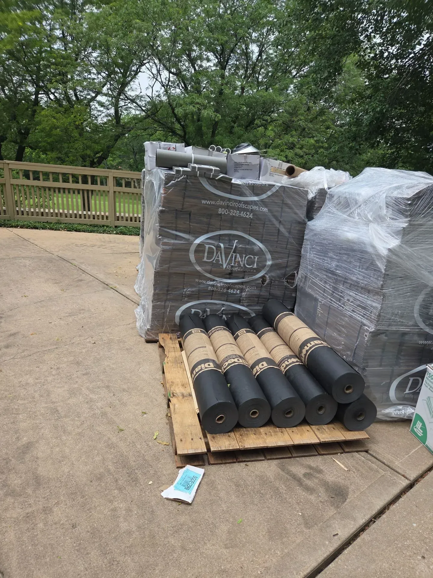 Pallet of roofing materials including rolled and stacked items, outside on a concrete surface.