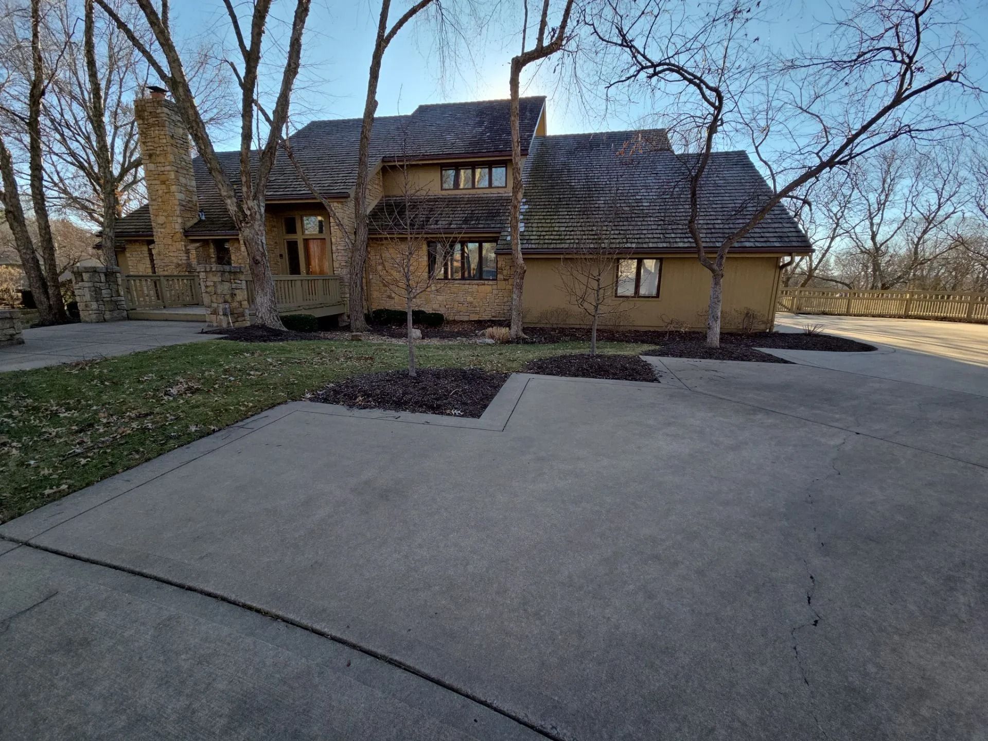 A two-story brown house with a concrete driveway and bare trees under a bright sky.