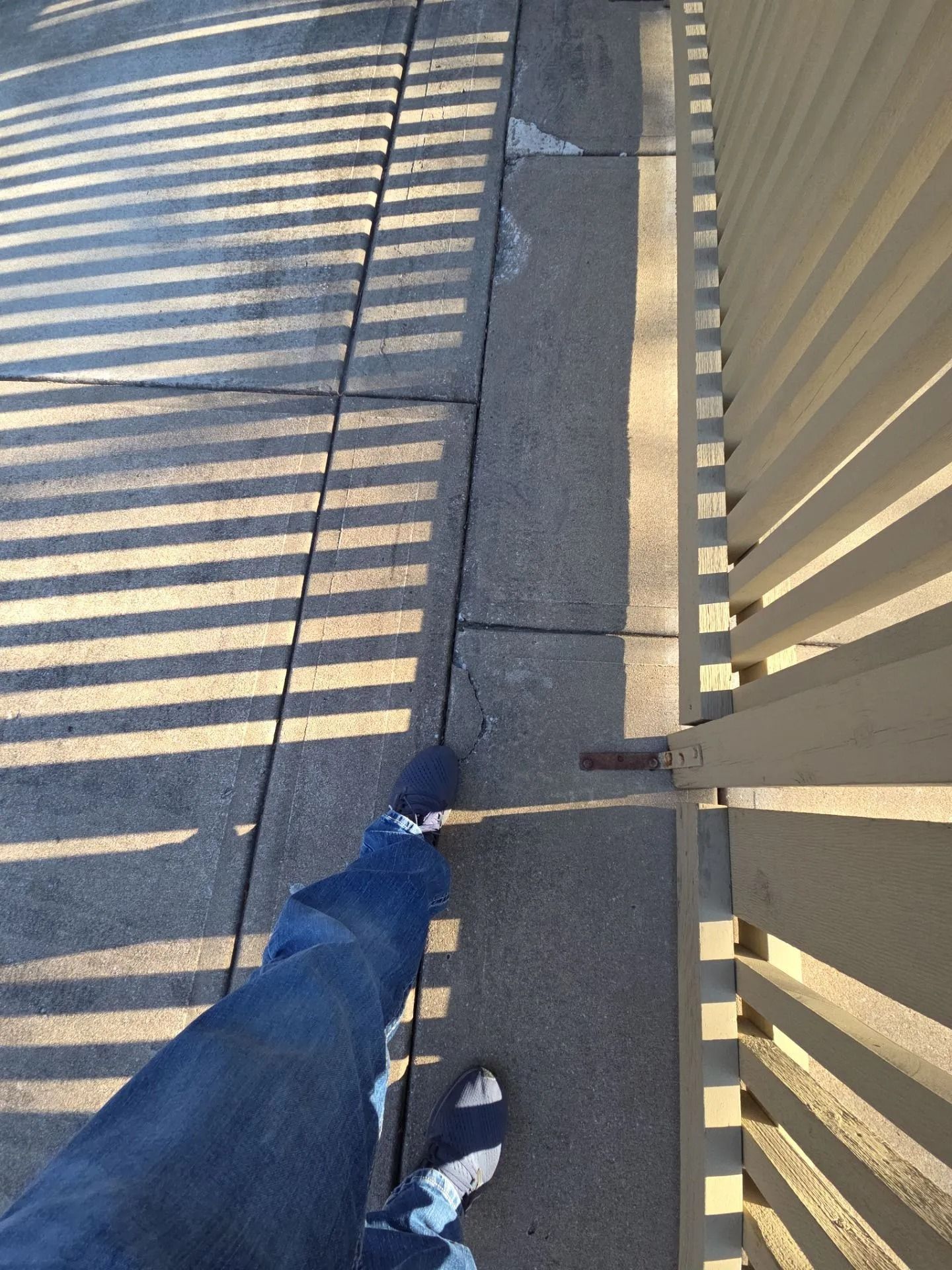 Person's legs in jeans and socks walking on sidewalk next to a fence casting striped shadows.