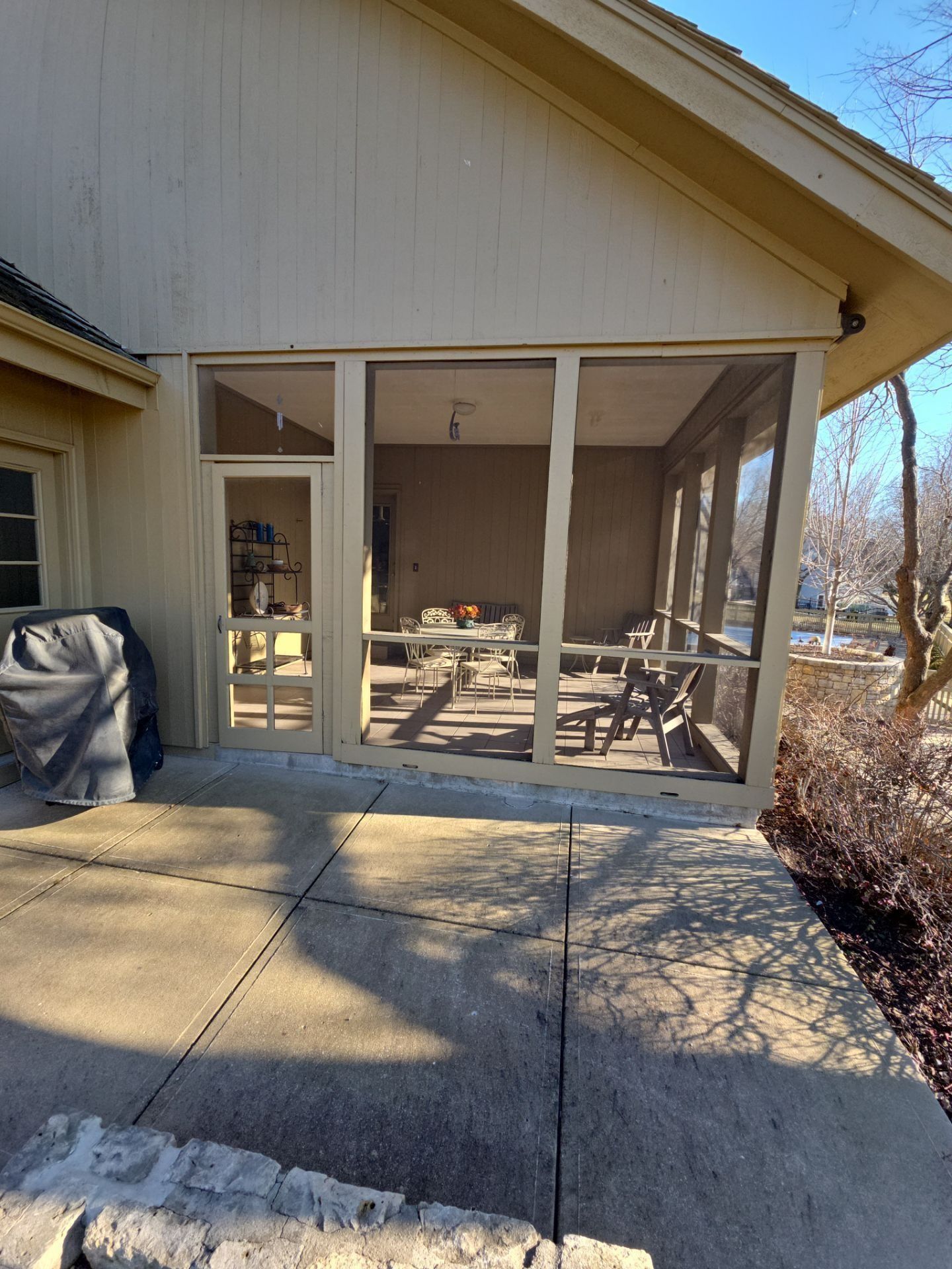 A screened porch with tan framing and screen walls, built onto a light-colored building, on a concrete patio.