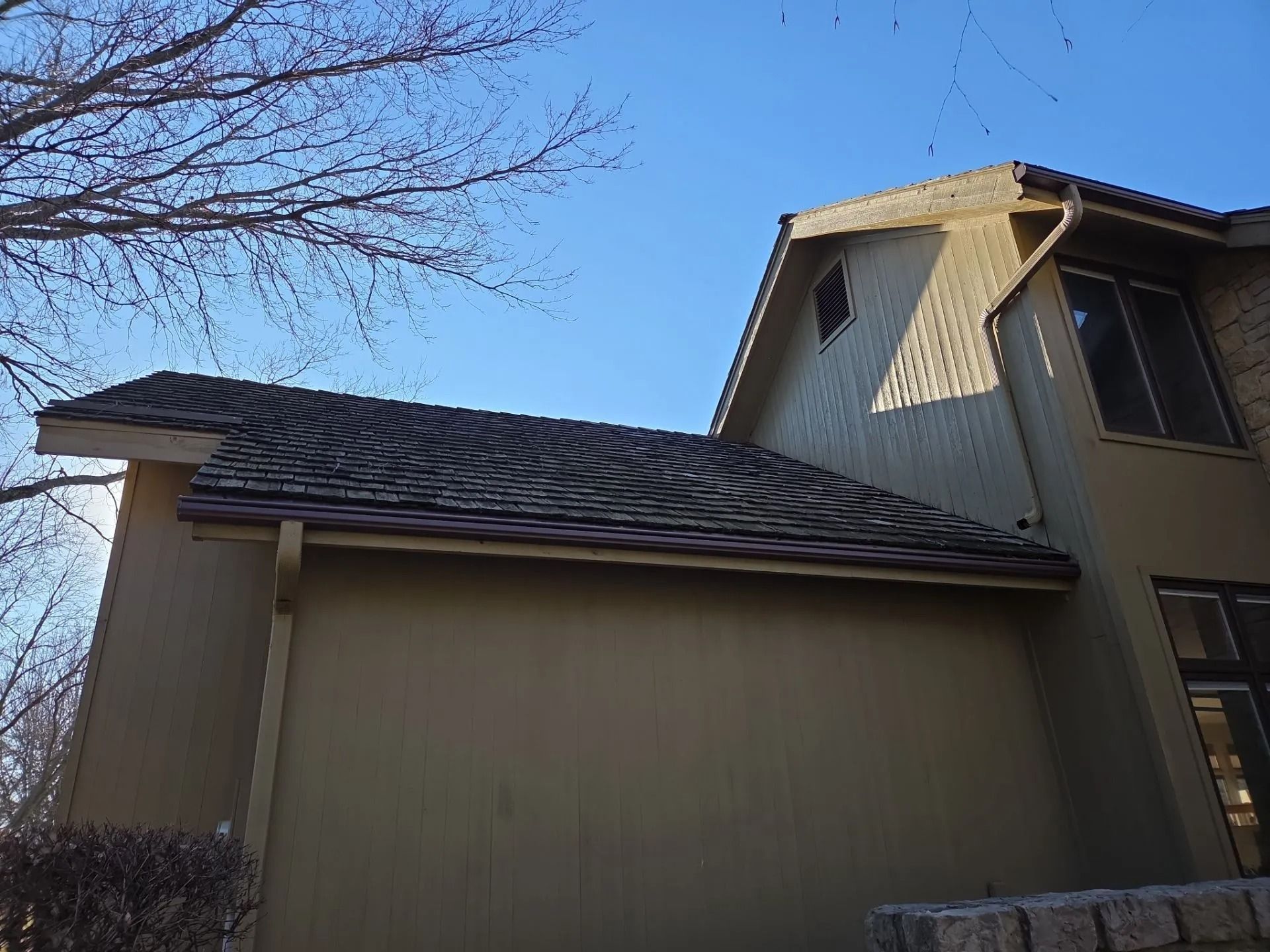 Tan house with dark roof and brown gutters against a clear blue sky.