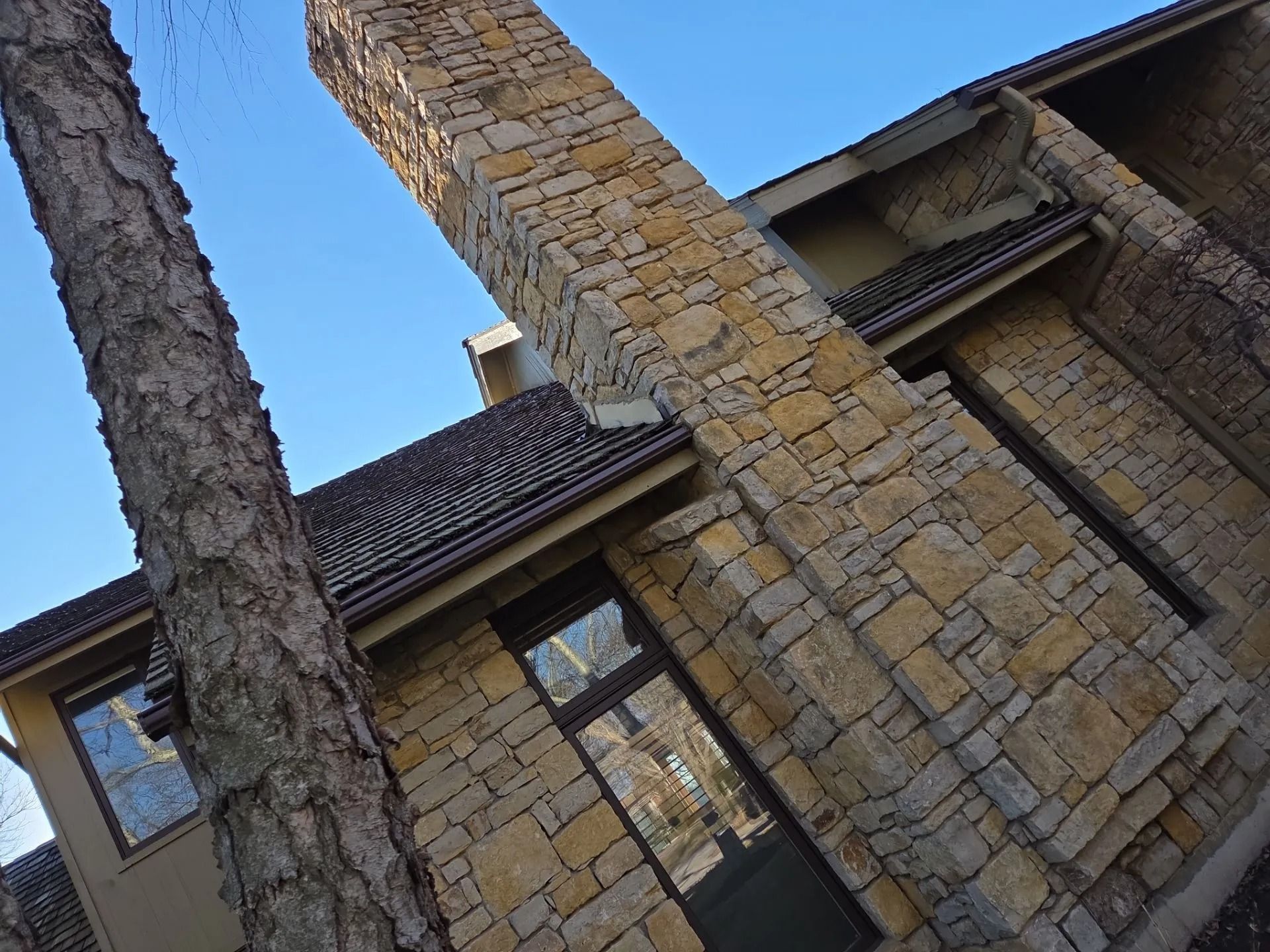 Stone-clad building with windows and chimney against a blue sky, tree in the foreground.