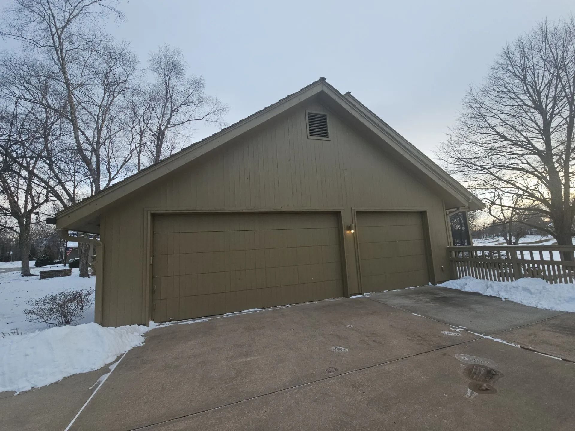 A two-car garage with brown doors, driveway, snow, and trees. Overcast sky.