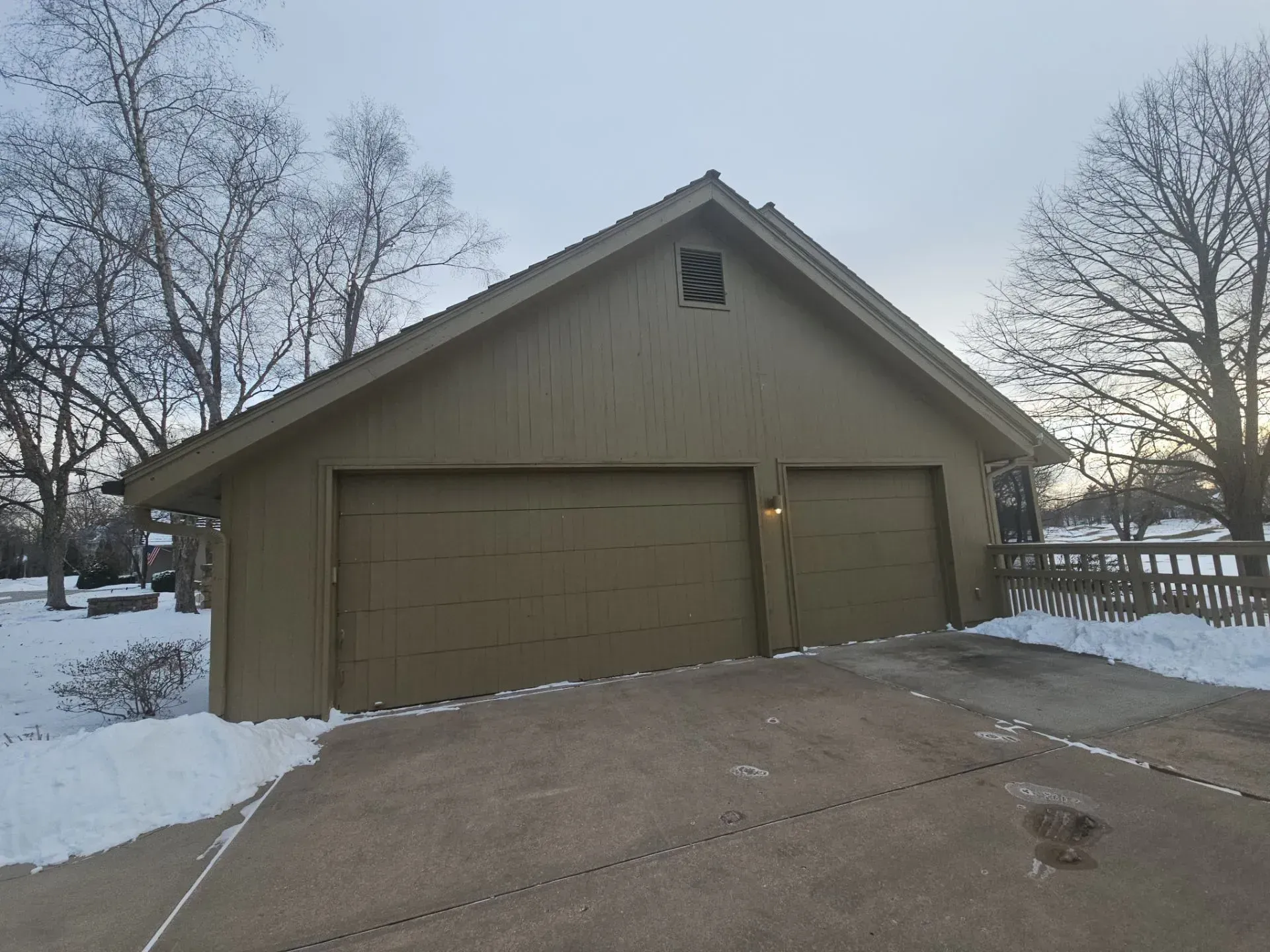 A two-car garage with brown doors and siding, a snowy driveway, and bare trees under a cloudy sky.