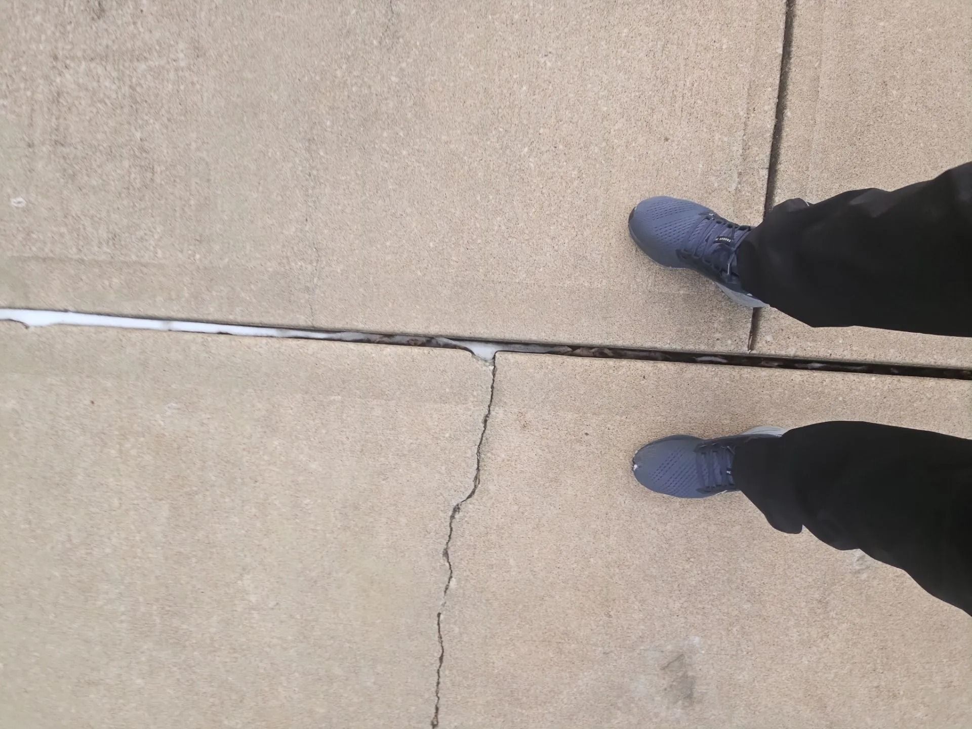 Feet in blue shoes on a concrete sidewalk, standing near a crack with possible ice.