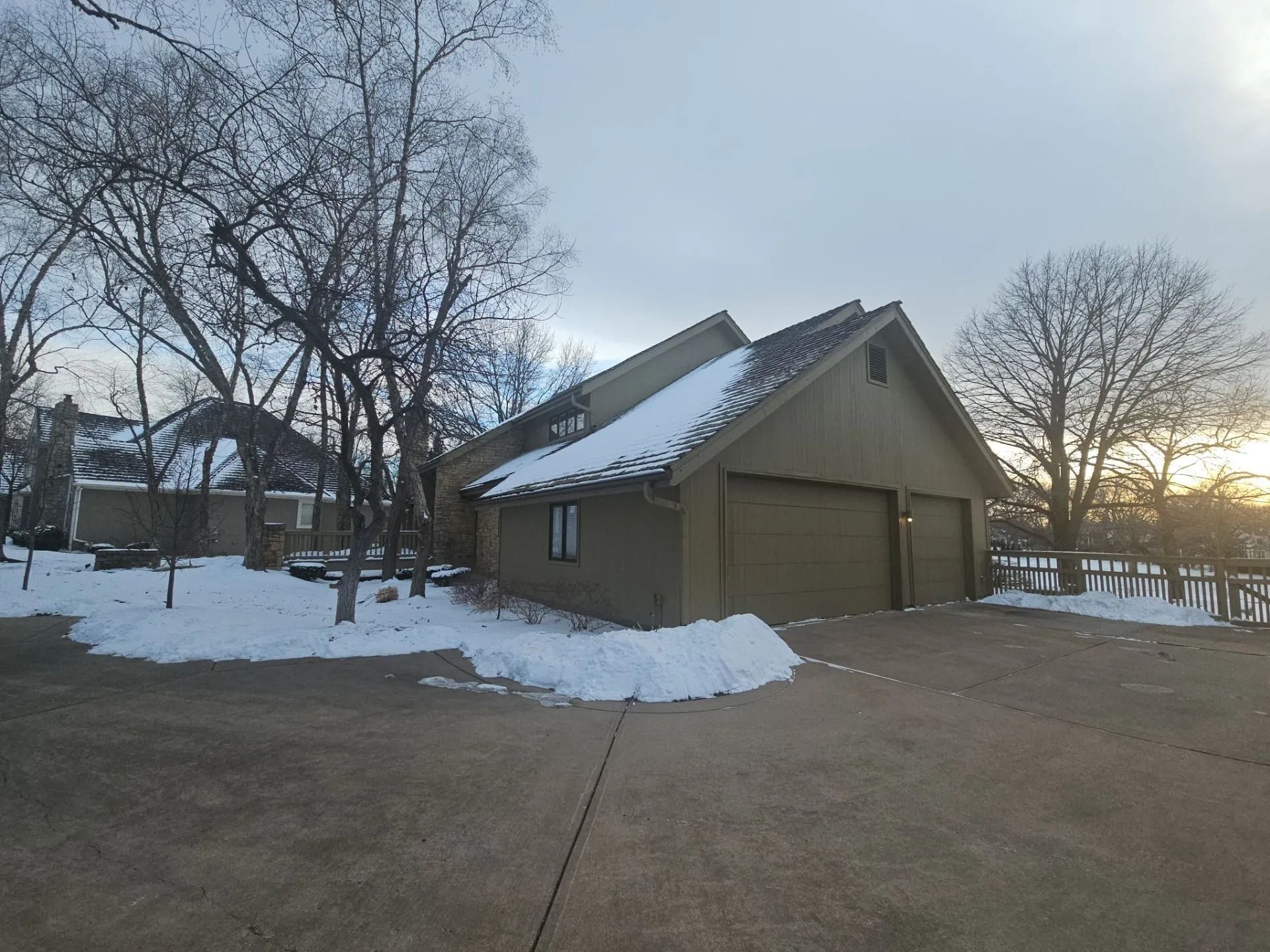 A house with a two-car garage covered in snow, bare trees, and a driveway on a cloudy day.