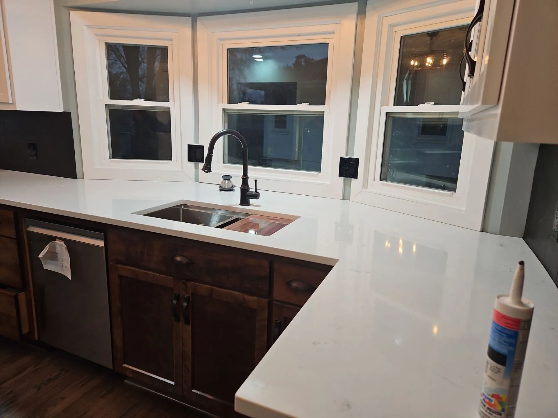 Kitchen with white countertops, dark wood cabinets, sink under windows, and caulk tube on the counter.