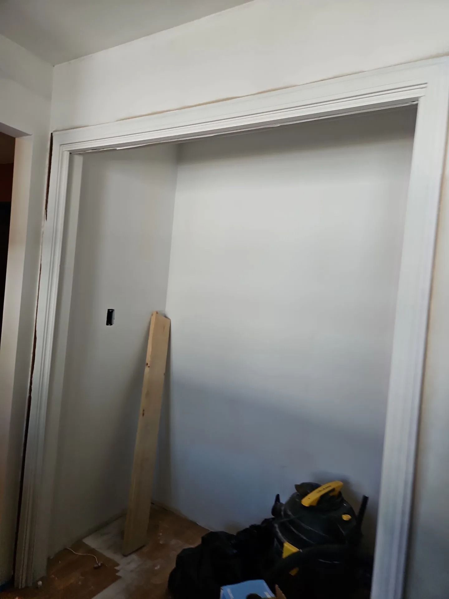 Empty closet with white walls and trim, construction in progress. A board leans against the wall.
